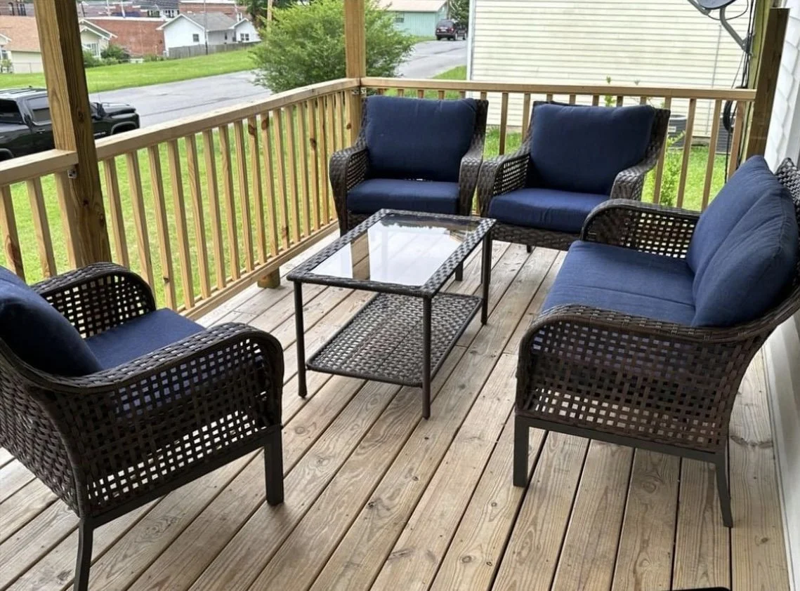 A wooden porch with four dark wicker chairs with blue cushions and a small glass-top table. Green grass and houses visible in the background.