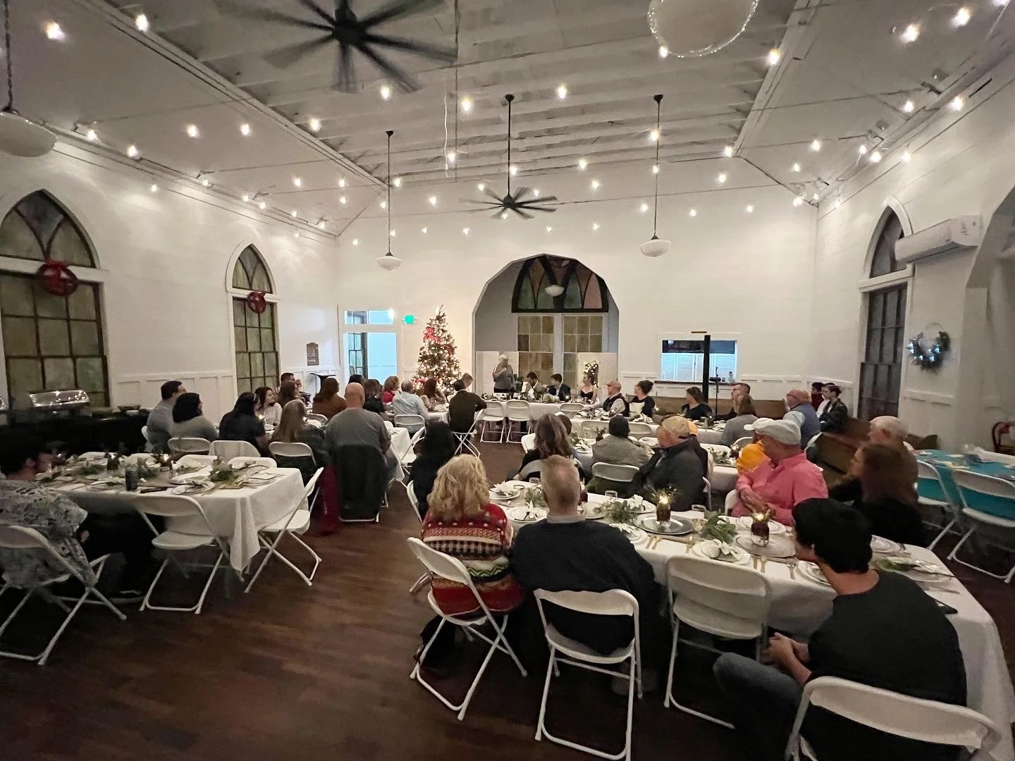 People seated at long tables in a decorated banquet hall, attending a holiday gathering with Christmas decorations, including a decorated Christmas tree and wreaths, and warm ambient lighting.