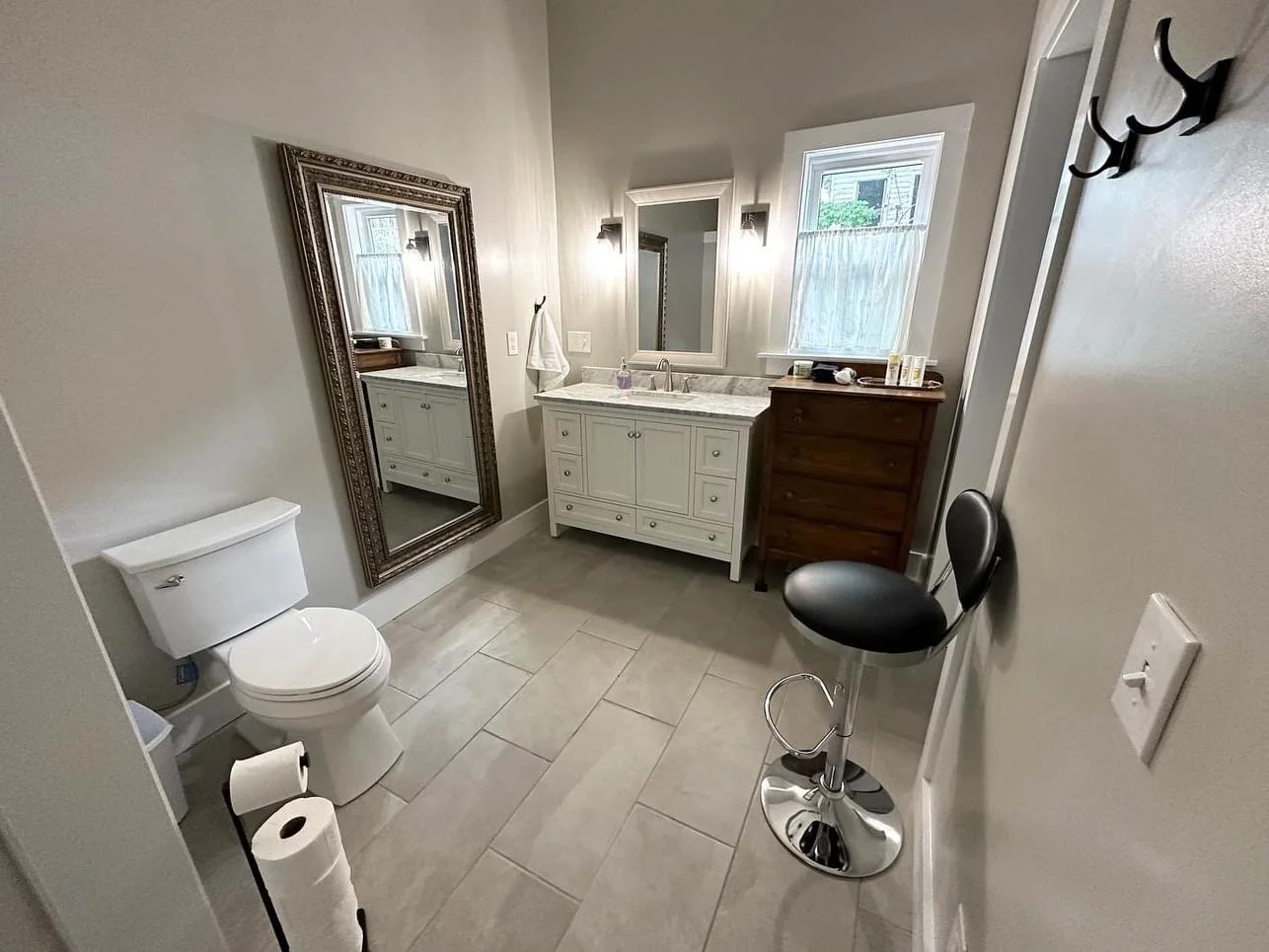 A small bathroom with a toilet, a white vanity with a marble countertop, a mirror, and a window with a curtain. There is a wooden chest of drawers, a black and silver barstool, a wall hook, and bathroom supplies on the countertop.