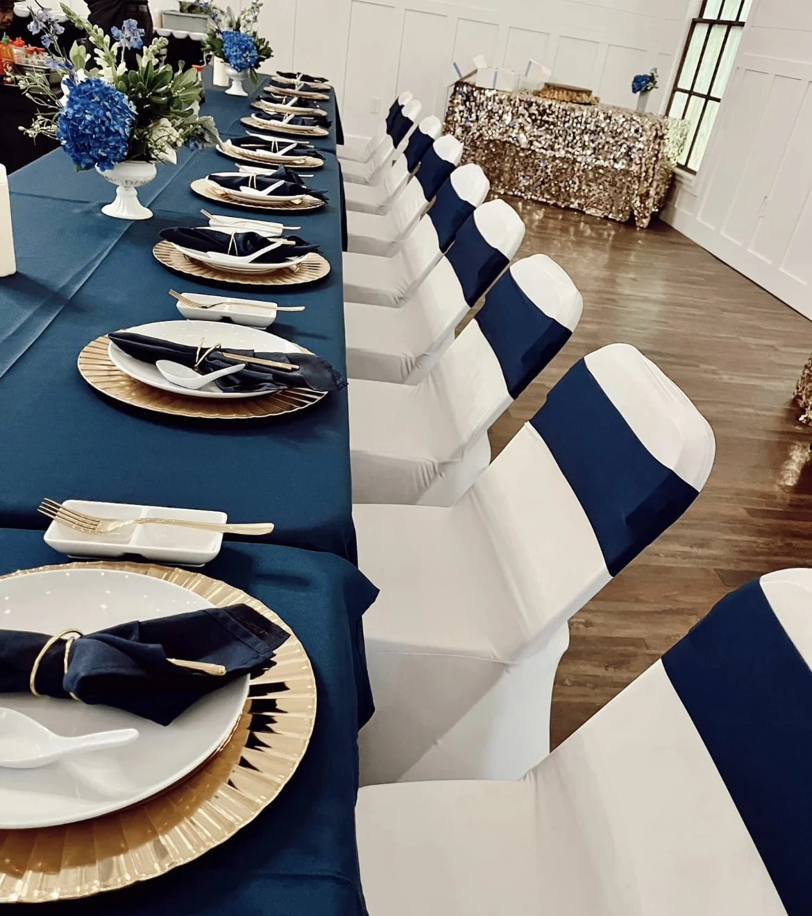 A long dining table set for a special event with blue tablecloths, gold-rimmed plates, black napkins wrapped with gold napkin rings, white serving spoons, and floral centerpieces of blue and white flowers.