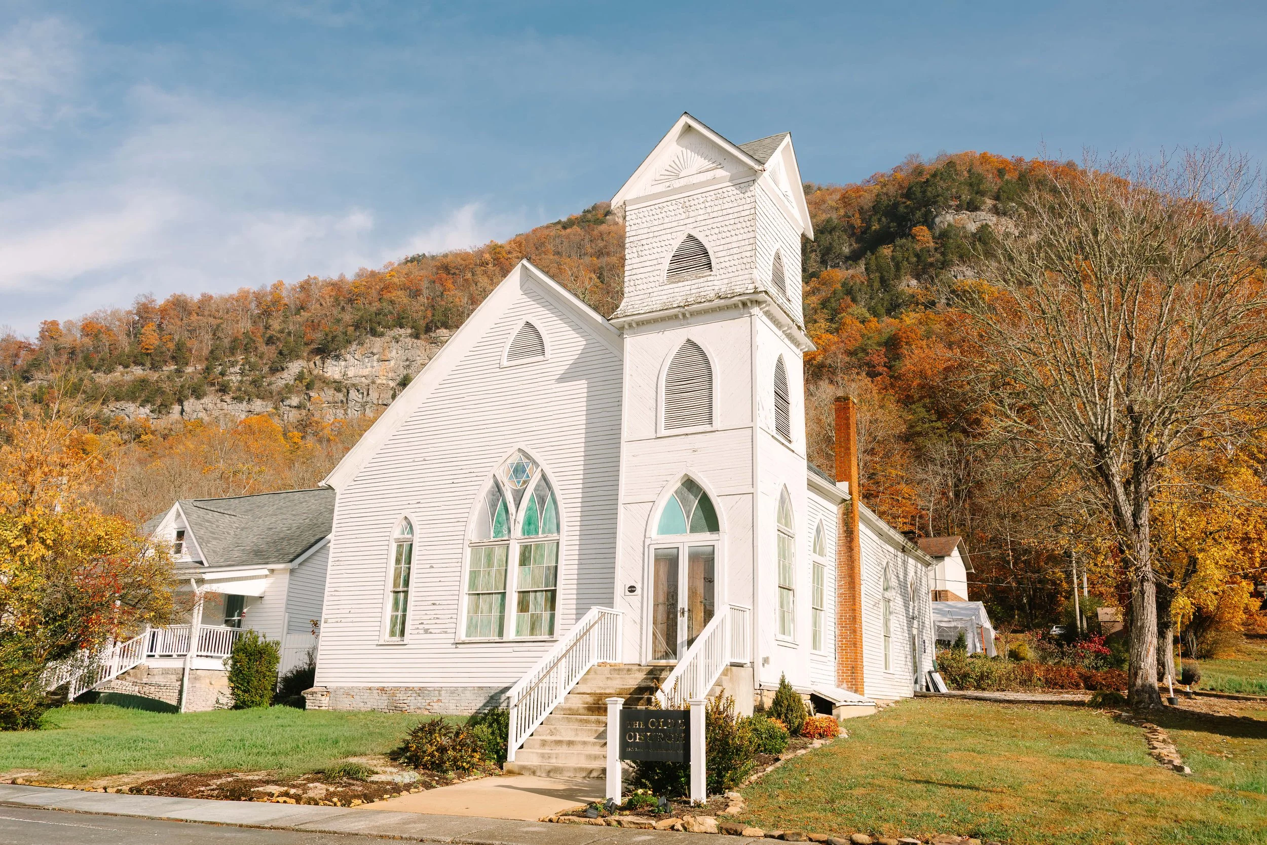 White wooden church with steeple and stained glass windows, set against a backdrop of fall foliage and a hillside, under a partly cloudy sky.