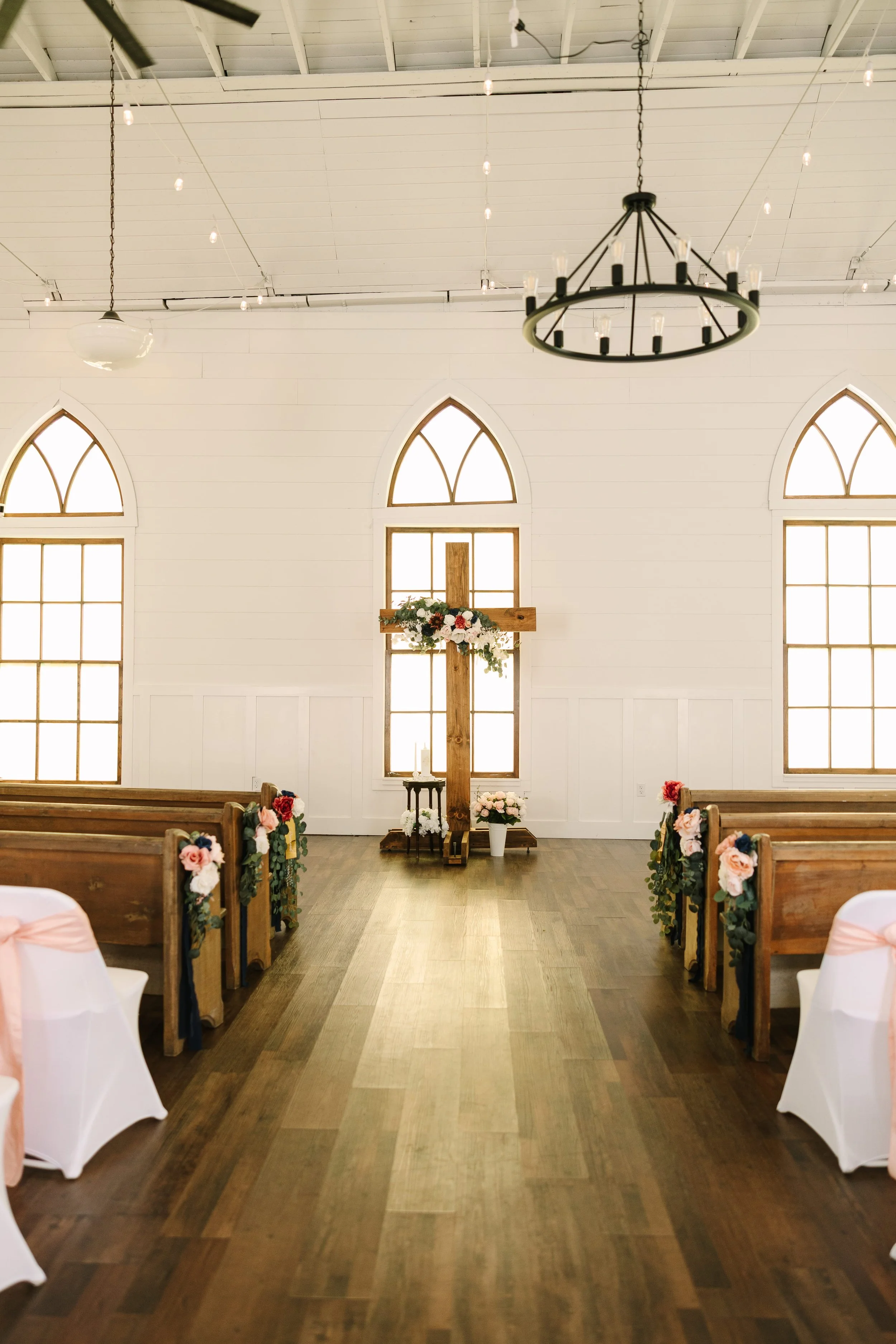Interior of a church or wedding venue decorated for a ceremony, with wooden pews adorned with flowers, arched windows, and a cross at the front, featuring a floral arrangement on the cross.