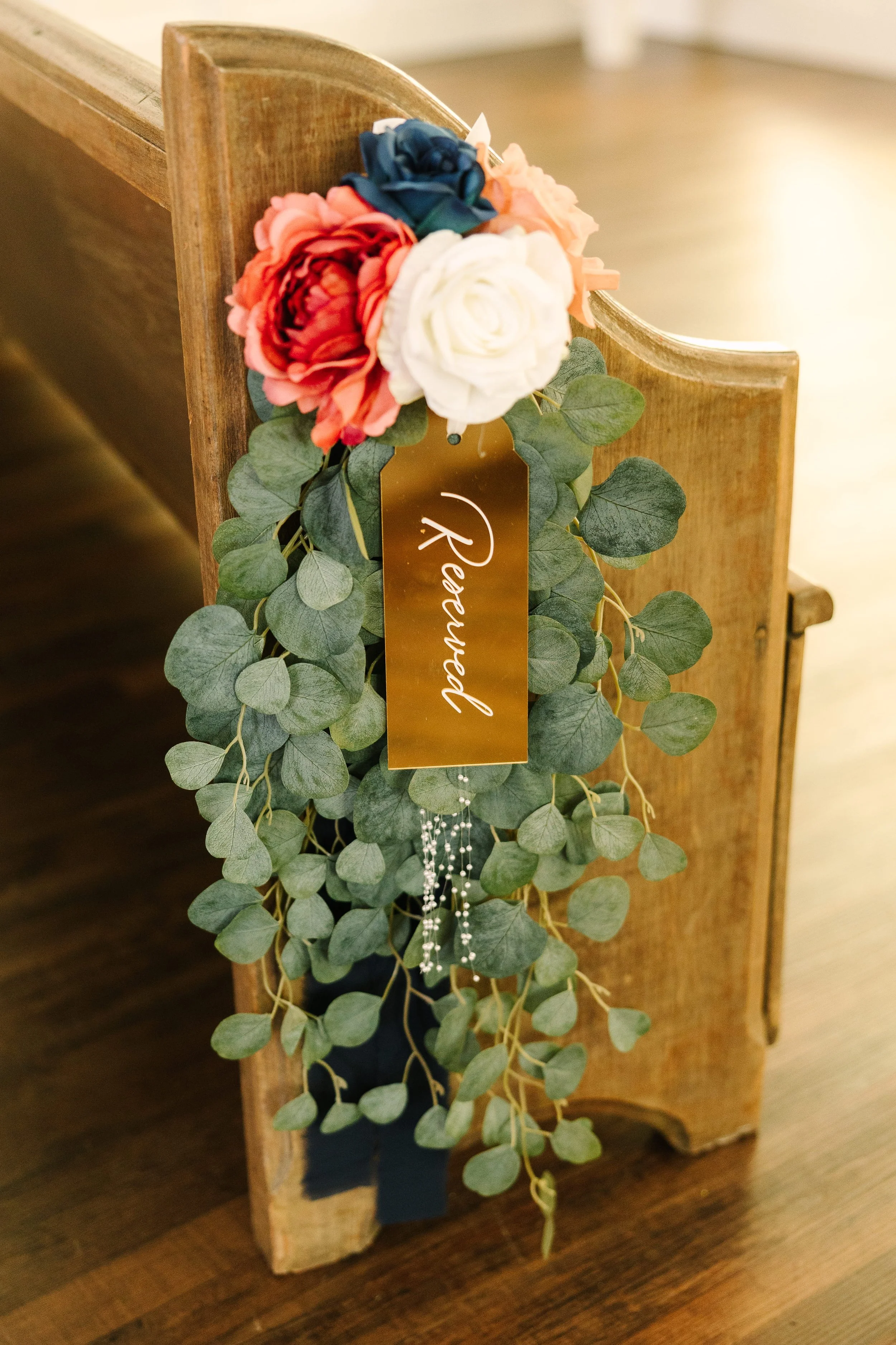 A floral decoration with yellow, pink, white, and blue artificial flowers, greenery, and a gold tag with the word 'Remember' attached to a wooden furniture piece.