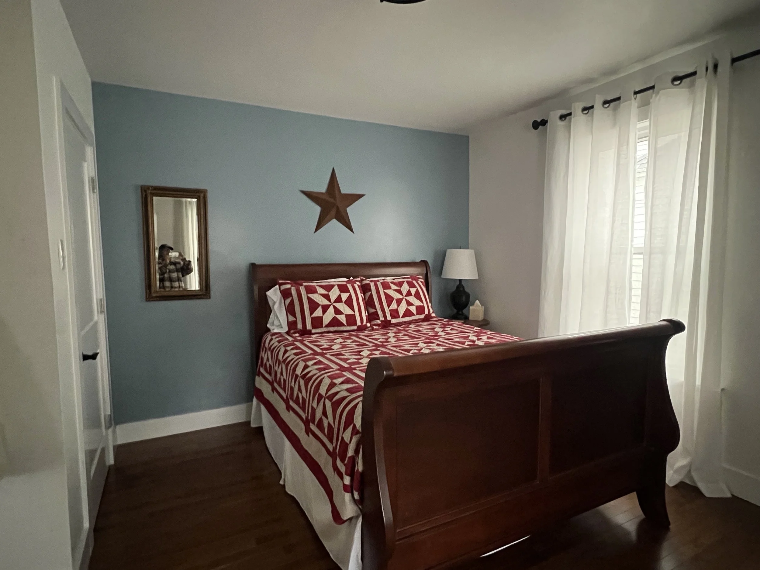 A bedroom with a wooden bed frame, red and white geometric patterned bedding, a white nightstand with a black lamp and tissue box, a wall mirror, a decorative star on the wall, and white curtains covering a window.