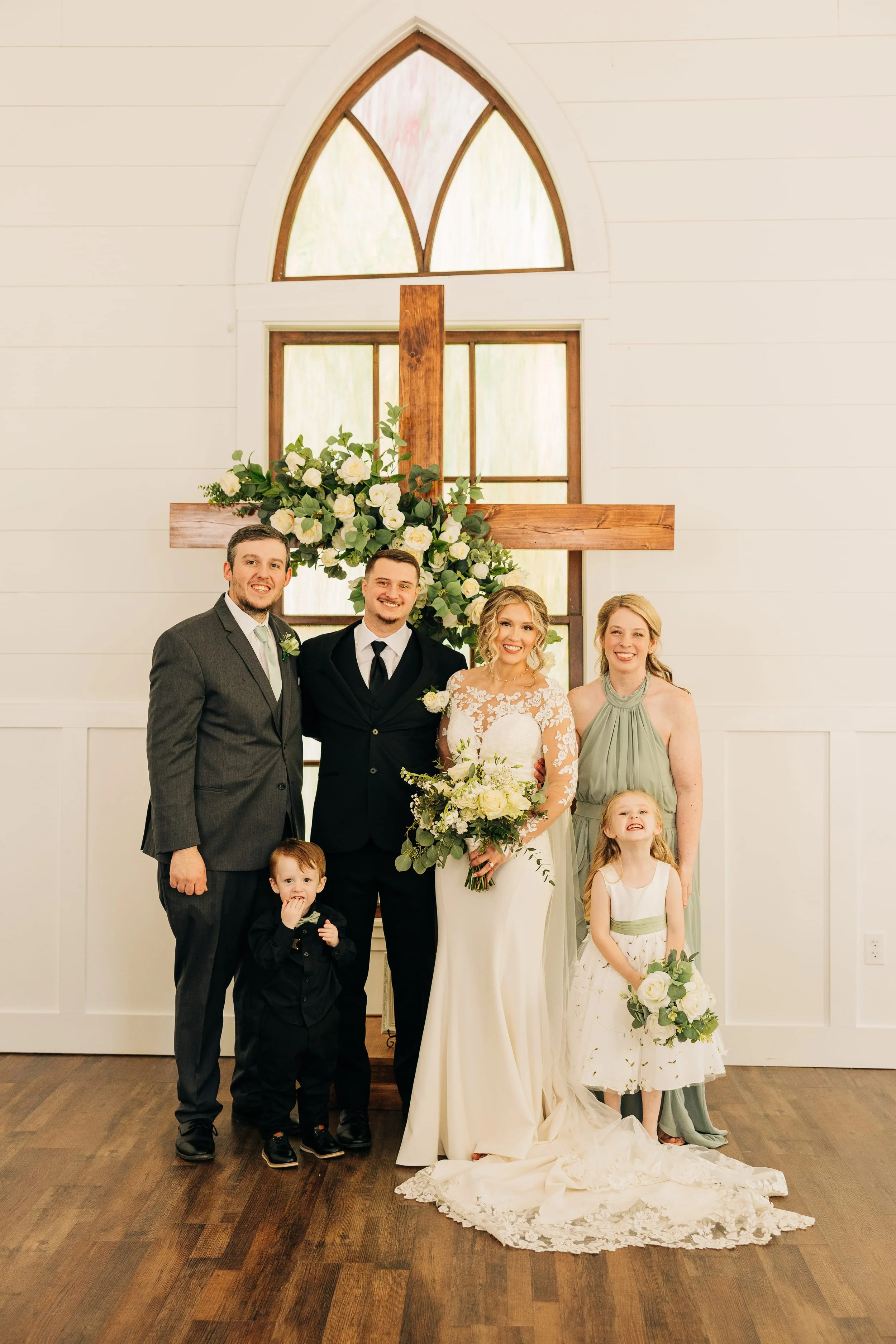 A wedding group portrait inside a church with a wooden cross decorated with white roses and greenery behind them. The bride is in a white lace and satin gown, holding a bouquet of white roses. The groom and three other adults, two women and one man, 