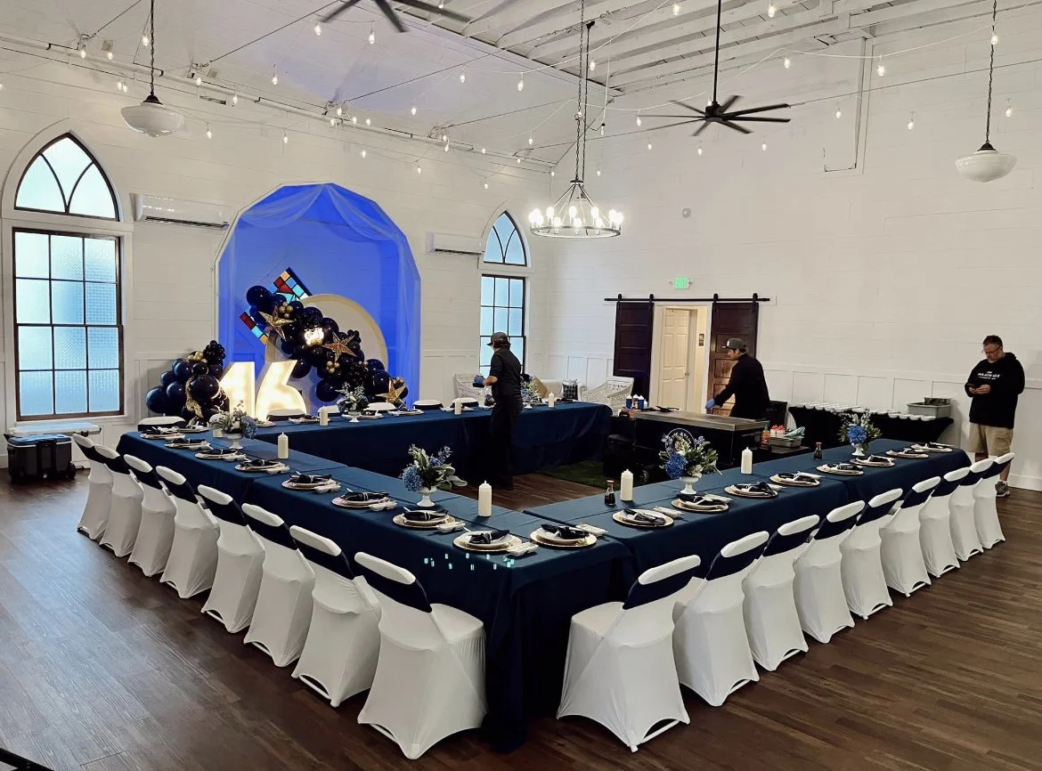 Event space with black and white table settings, white chair covers with black sashes, and a decorated head table with balloons, a Rubik's Cube, and star and moon decorations. Multiple light strings hang from the ceiling, and there are two men prepar