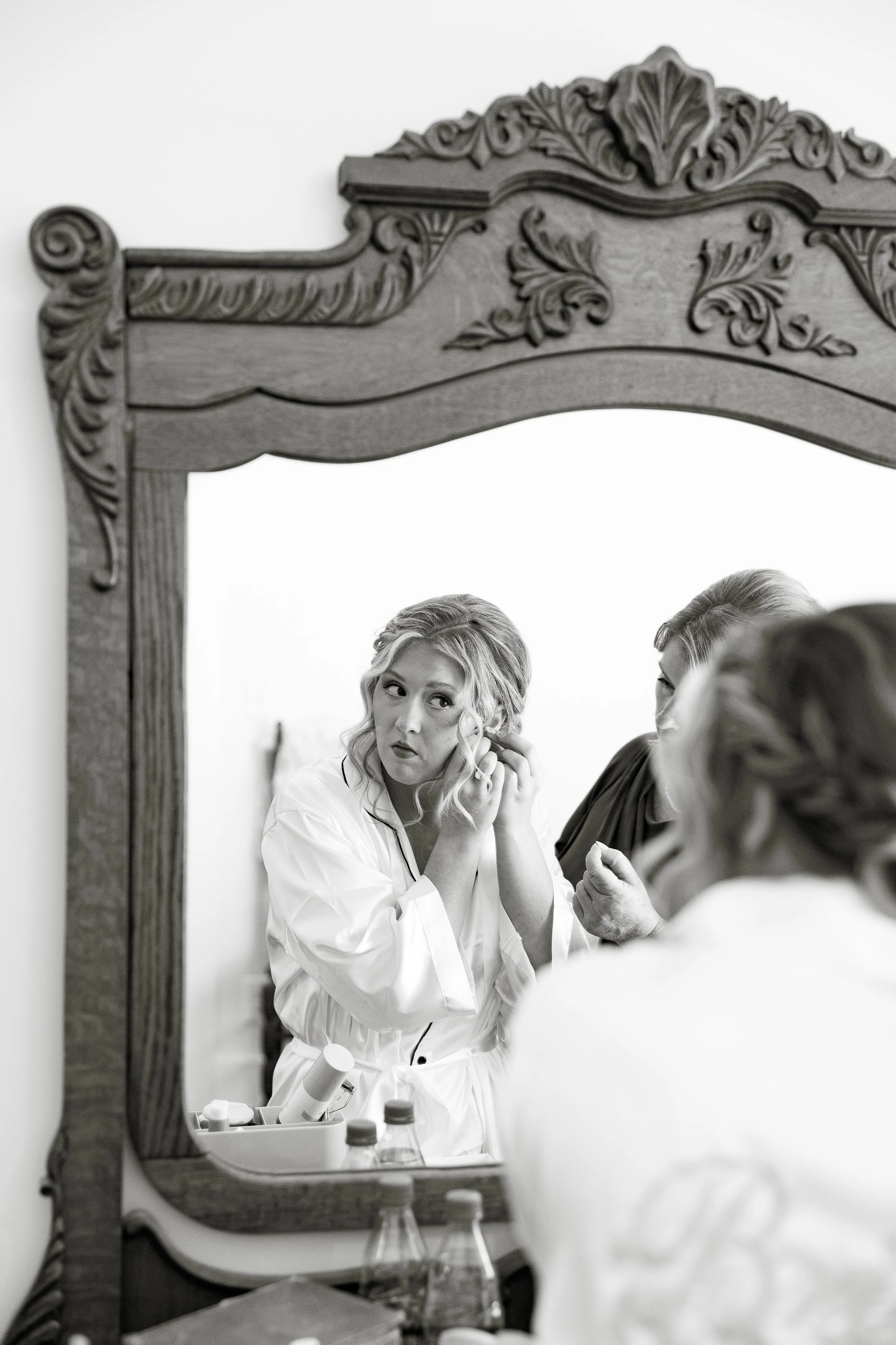 A woman is looking at herself in a mirror, adjusting her earring while styling her hair. The mirror has decorative wood carvings on the top. There is a vanity table below with bottles and a makeup tray.