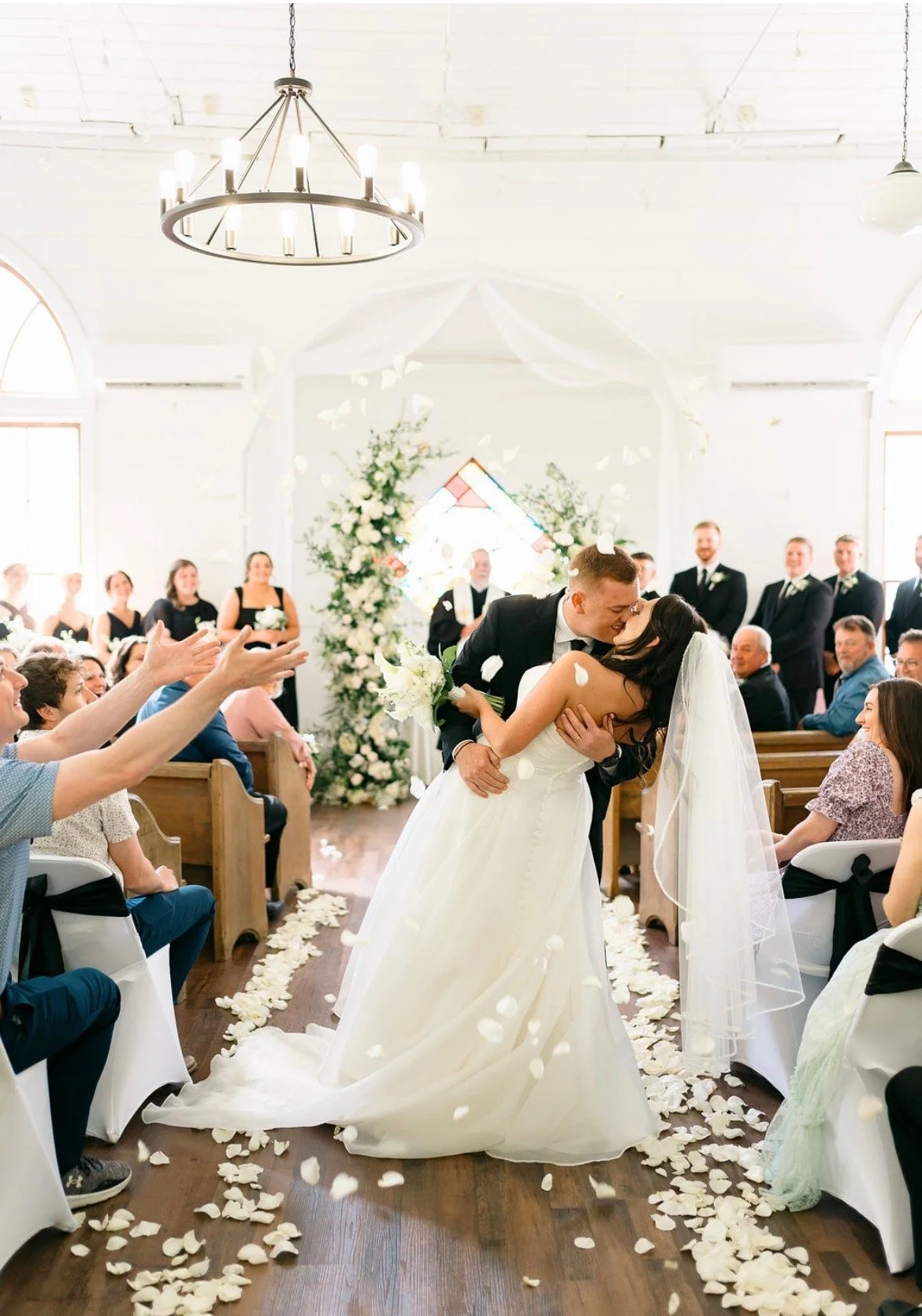 A wedding ceremony inside a church with bride and groom kissing, surrounded by friends and family. Rose petals fall from above, and floral arrangements decorate the background.