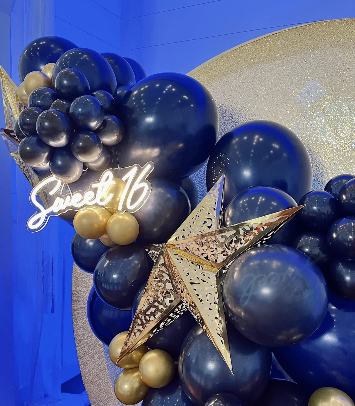 Balloon decoration with dark blue and gold balloons, a gold star, and a neon sign that reads 'Sweet 16' for a celebration.