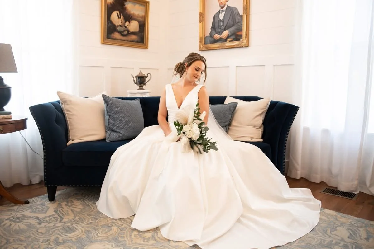 A bride in a white wedding gown sitting on a dark blue couch, holding a bouquet of white roses and greenery, in a room with white walls, natural light, and framed artwork hanging above.