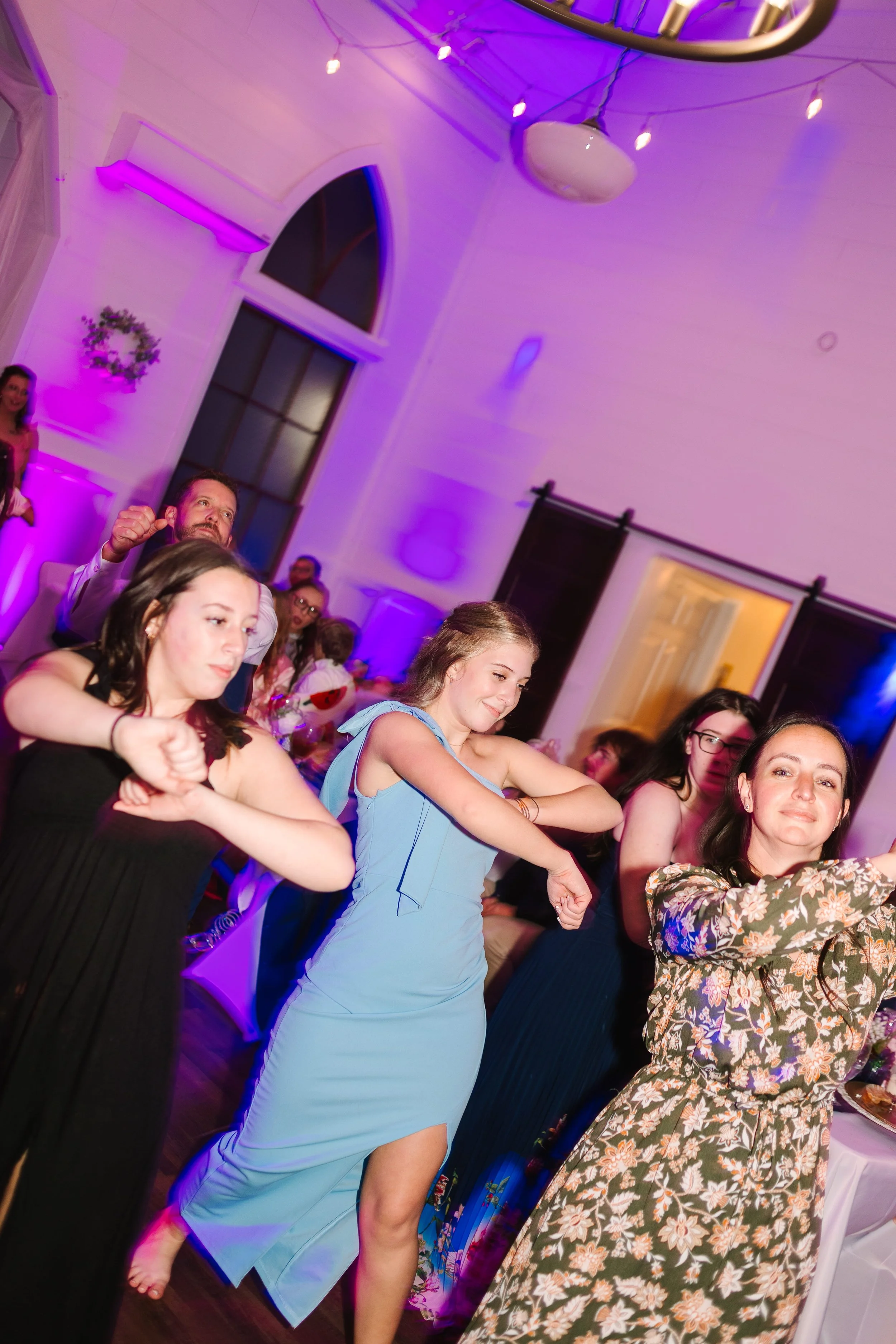 People dancing at a party with colorful purple and pink lighting, including women in dresses and a man with a beard, in a decorated indoor venue with large windows and string lights.