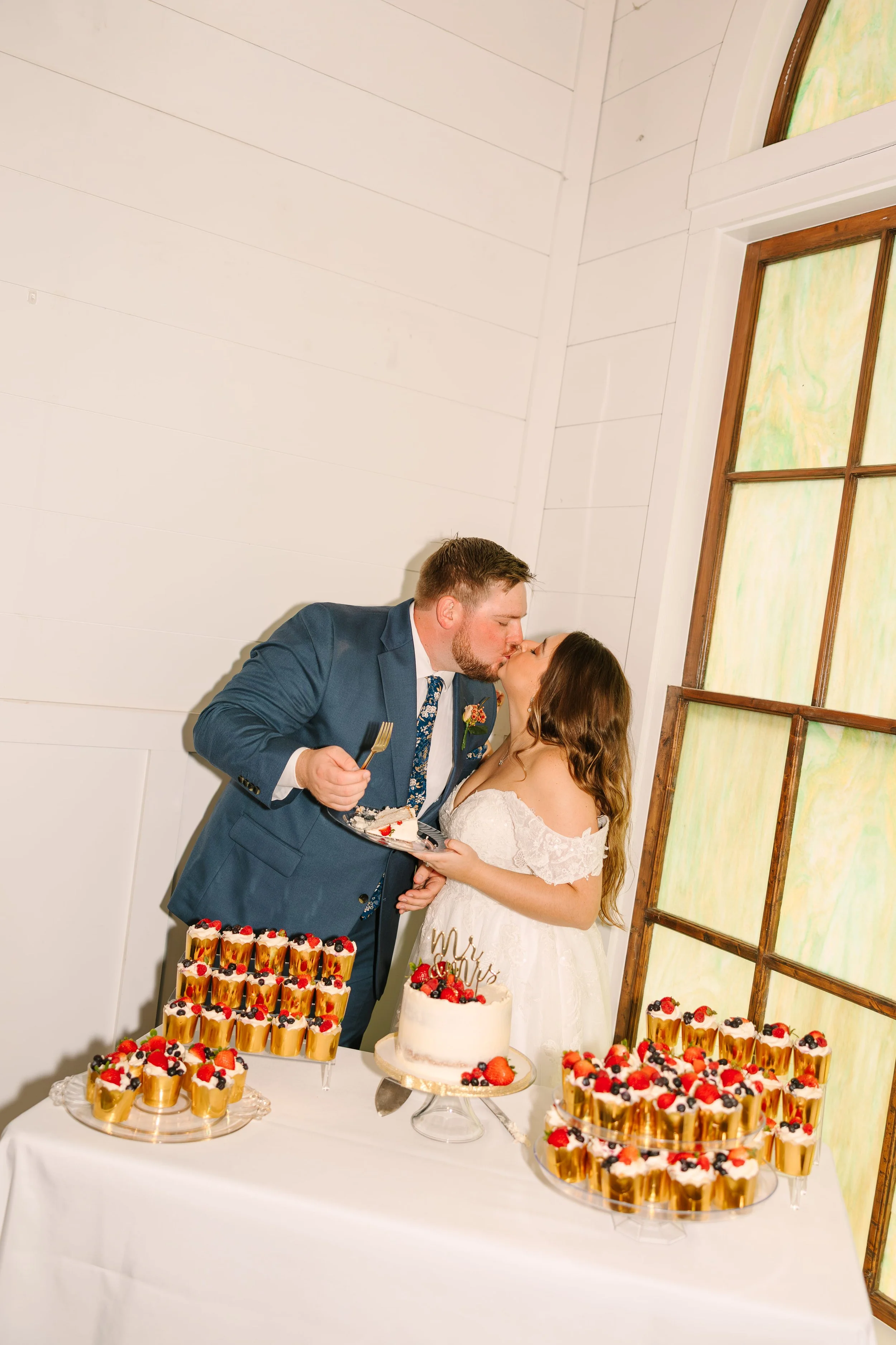 A newlywed couple sharing a kiss at their wedding reception. The groom is wearing a gray suit and the bride is in a white wedding dress holding a plate of cake. A table with a variety of desserts, including a wedding cake and individual parfaits topp