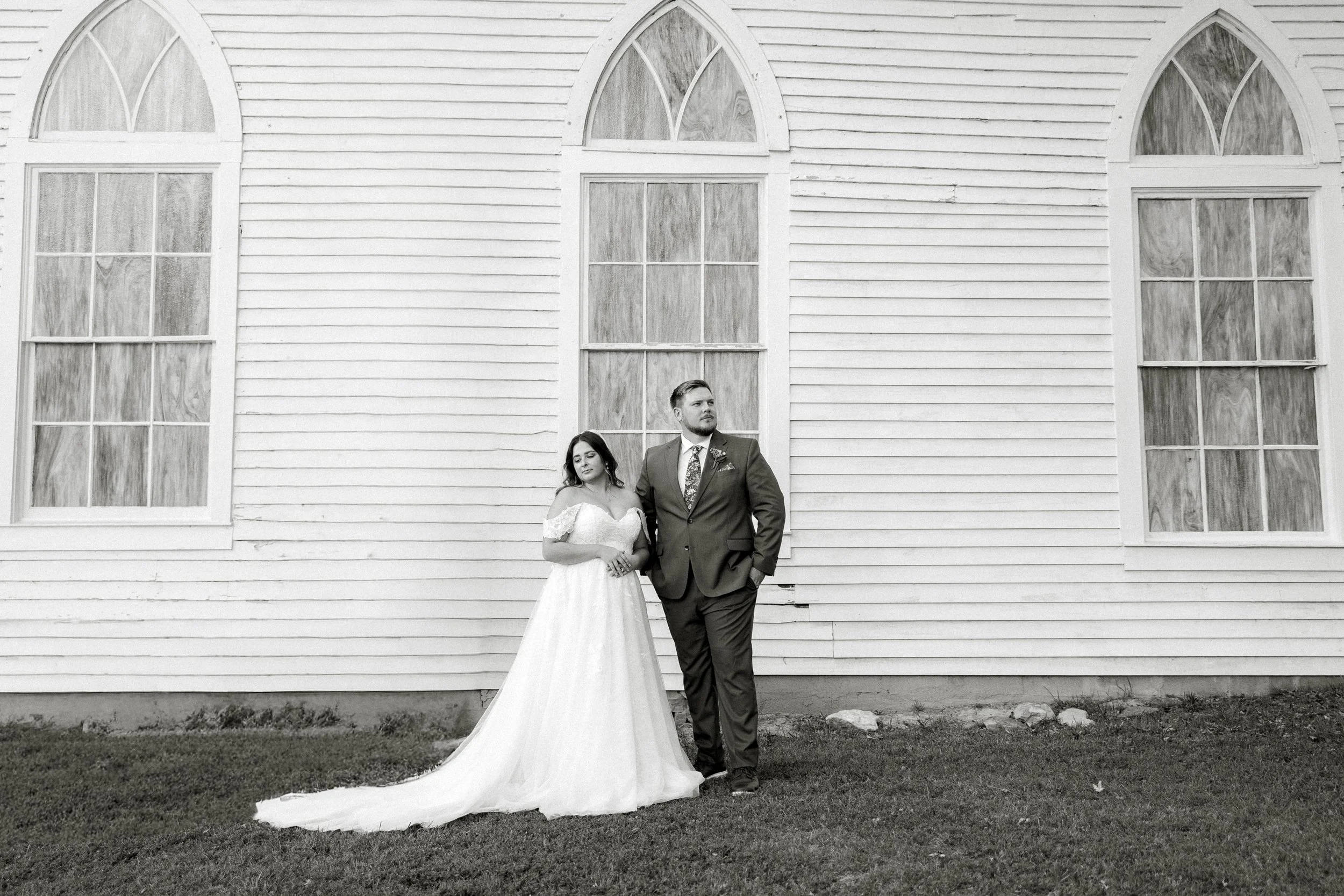 A newlywed couple stands in front of a white wooden church with large arched windows. The bride is wearing a white wedding gown, and the groom is in a dark suit with a patterned tie. They are looking away from each other, with the bride's eyes closed