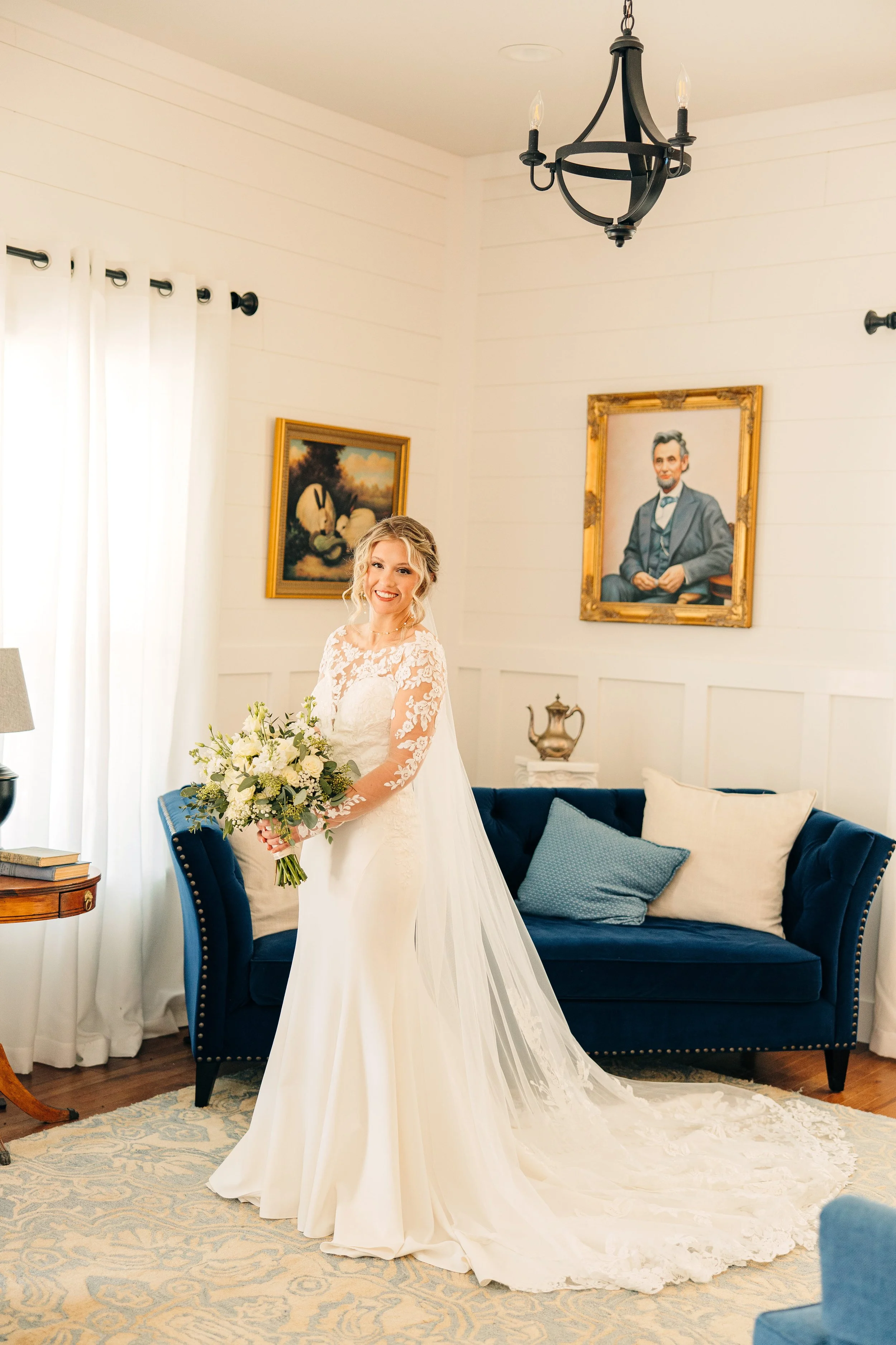 A smiling bride in a white wedding dress holding a bouquet of white flowers in a room with white panel walls, a dark blue sofa with blue and white pillows, and two framed paintings of rabbits and a man in a suit hanging on the walls.