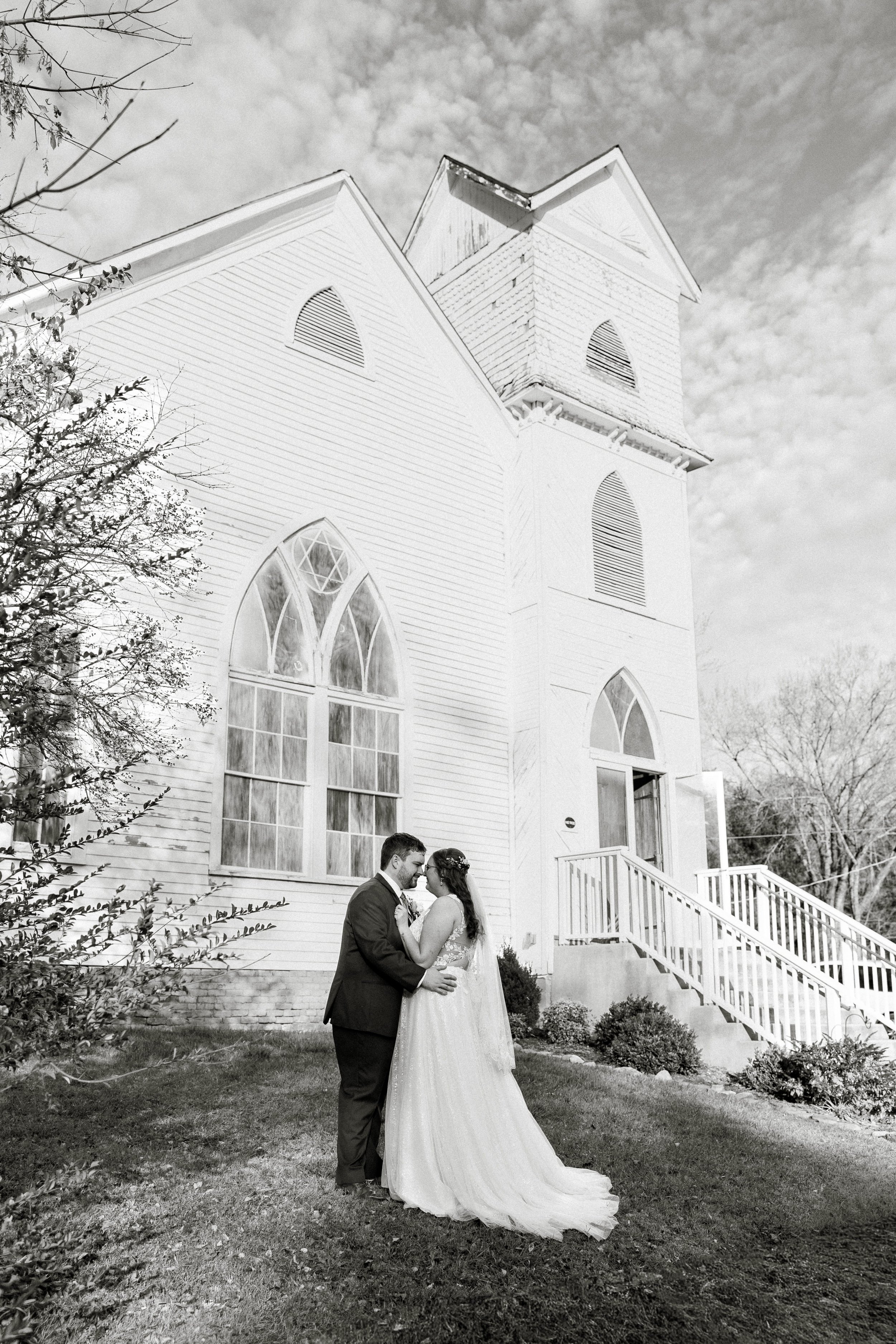 A black and white photo of a bride and groom holding hands and looking at each other in front of a white church with large arched windows and staircase.