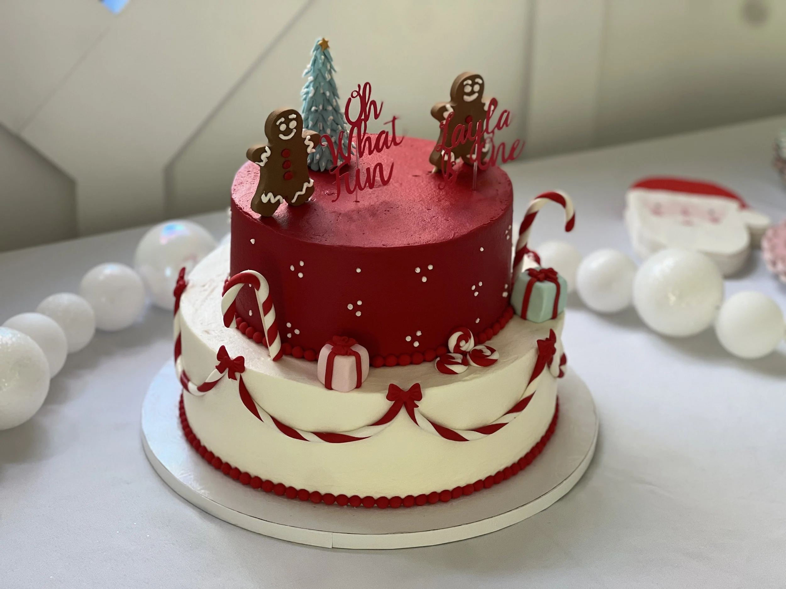 A two-tier Christmas cake decorated with red and white candy canes, red and white fondant gift boxes, small white and red stars, and white edible snow dots, topped with gingerbread cookies, a small blue Christmas tree, and a red cake topper with the 