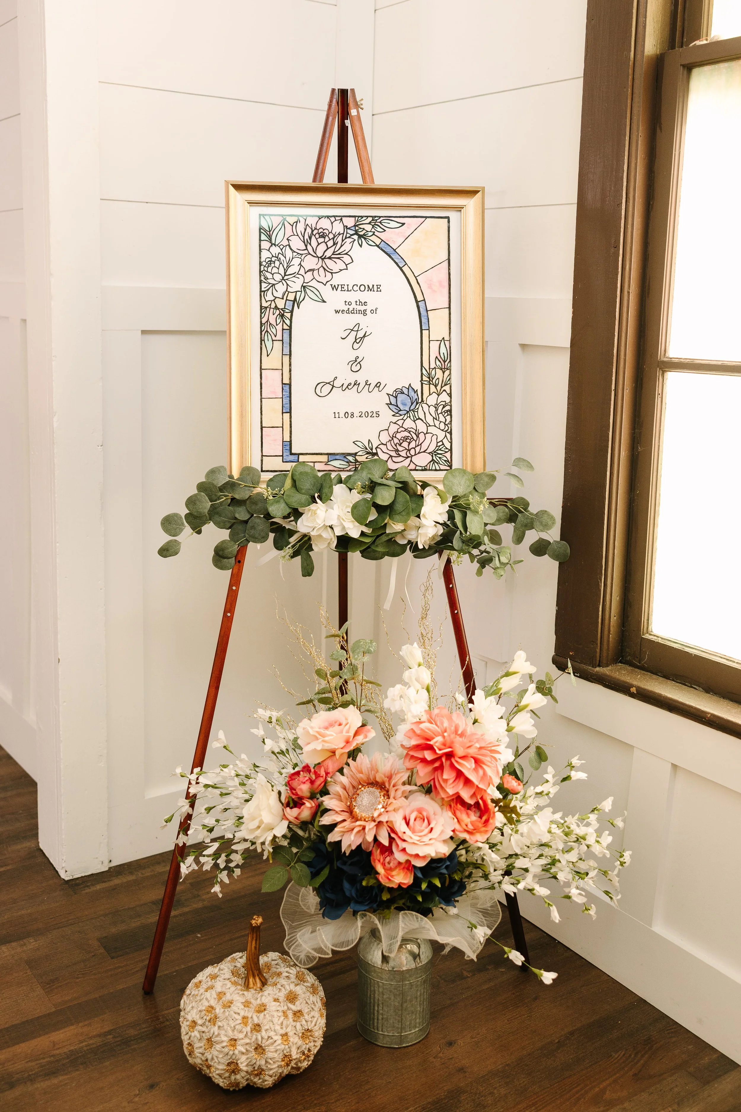 A wedding welcome sign on a wooden easel, decorated with flowers and greenery, displaying the names Ai and Sierra and the wedding date 11.08.2025, positioned next to a floral arrangement with pink, white, and peach flowers, and a decorative pillow on