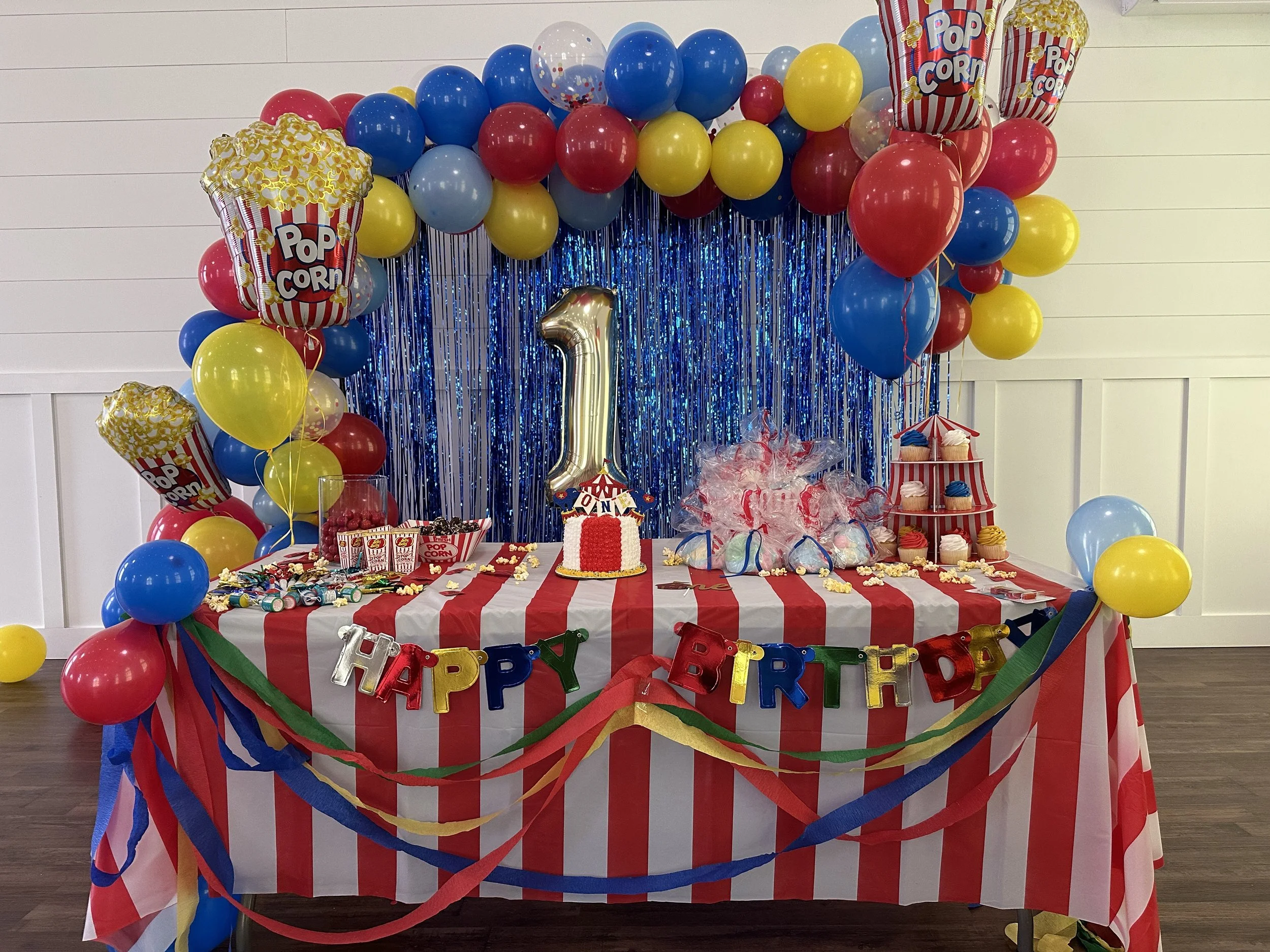 Colorful birthday party table decorated with red and white striped tablecloth, balloons in red, blue, yellow, and clear, blue metallic fringe backdrop, butterfly-shaped popcorn boxes, a large gold number one balloon, a birthday cake with a red and wh