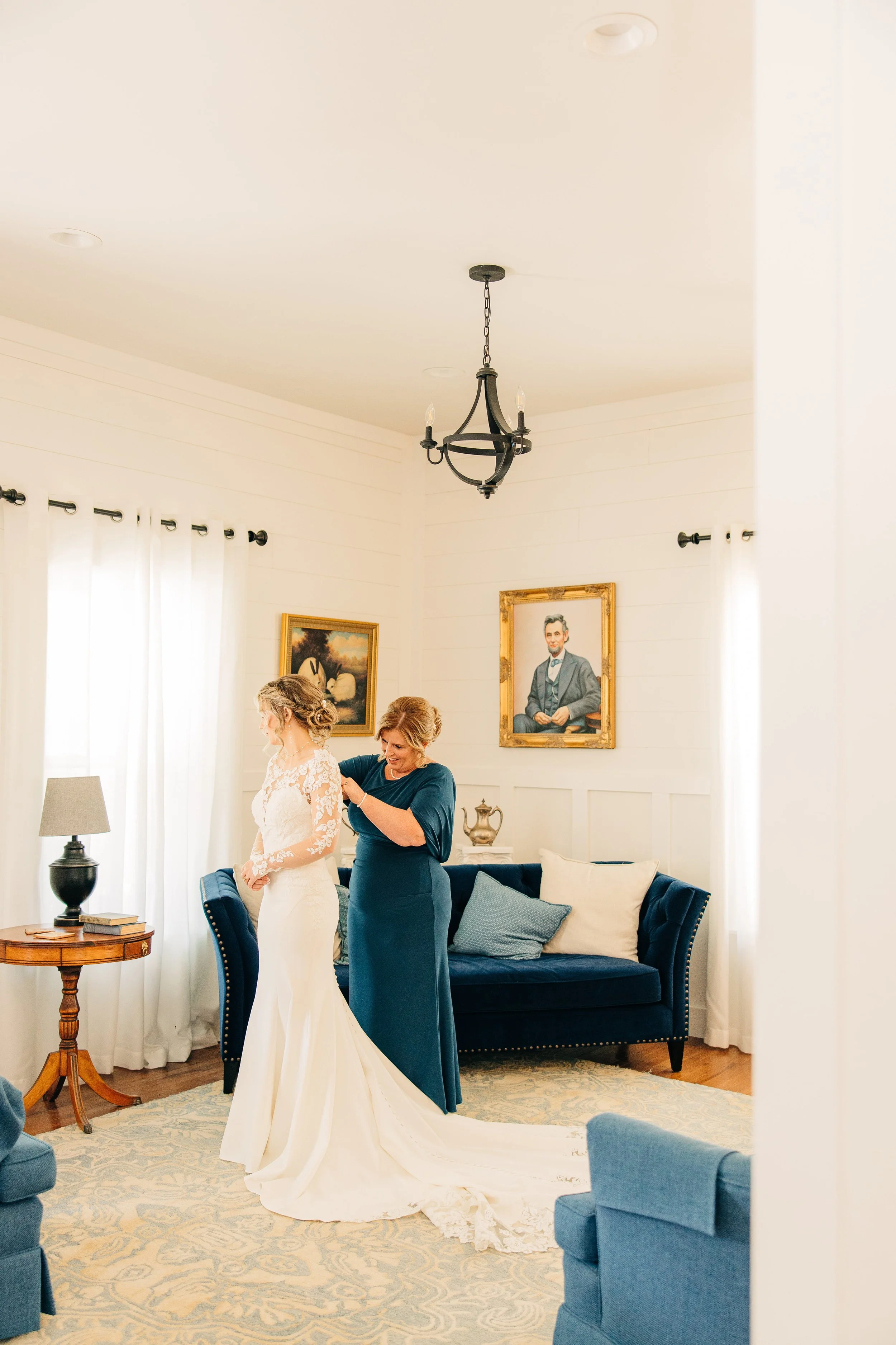 A bride in a white wedding dress stands in a living room while a woman in a blue dress adjusts her dress. Two portraits hang on the wall behind them, and there are large windows with white curtains. The room has a dark blue sofa with pillows, a wooden side table, and a patterned rug.