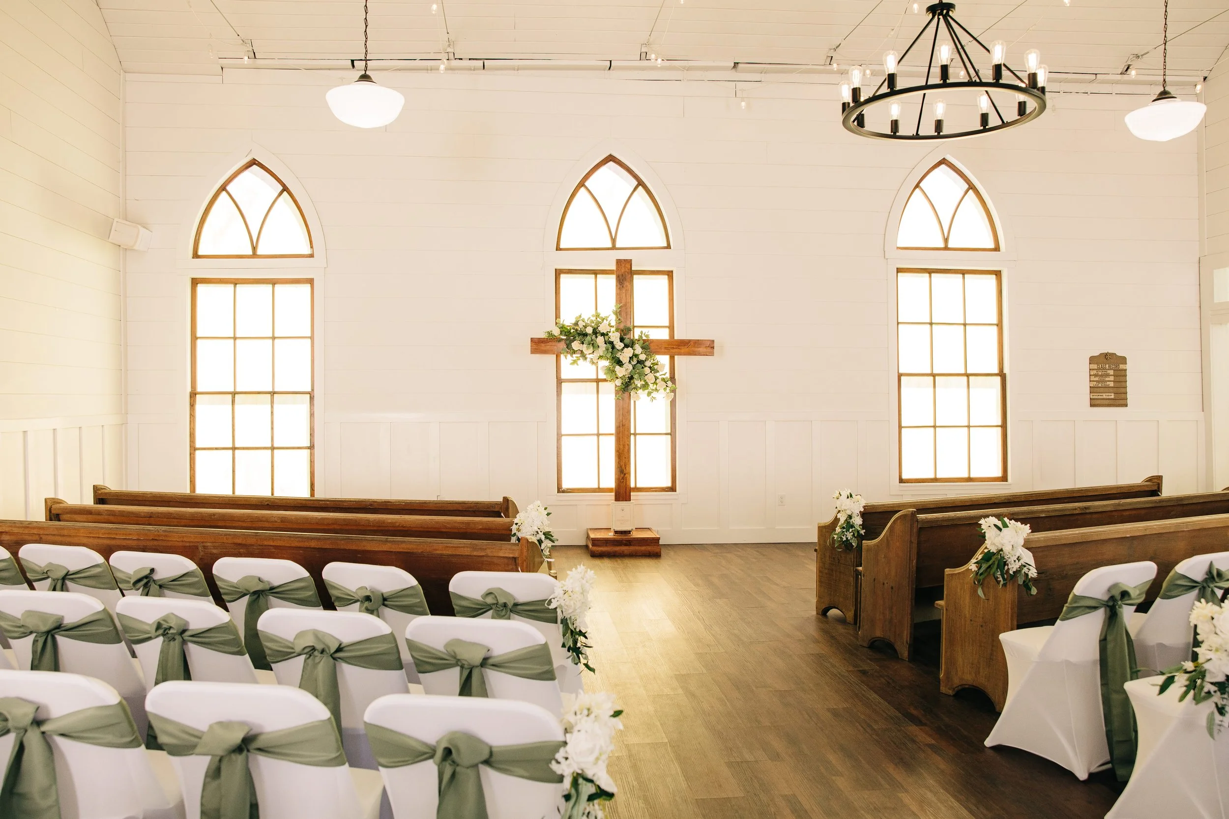 Interior of a small church decorated for a wedding with white chairs tied with green ribbons, a wooden altar with a cross and flower arrangements, and three tall arched windows letting in natural light.