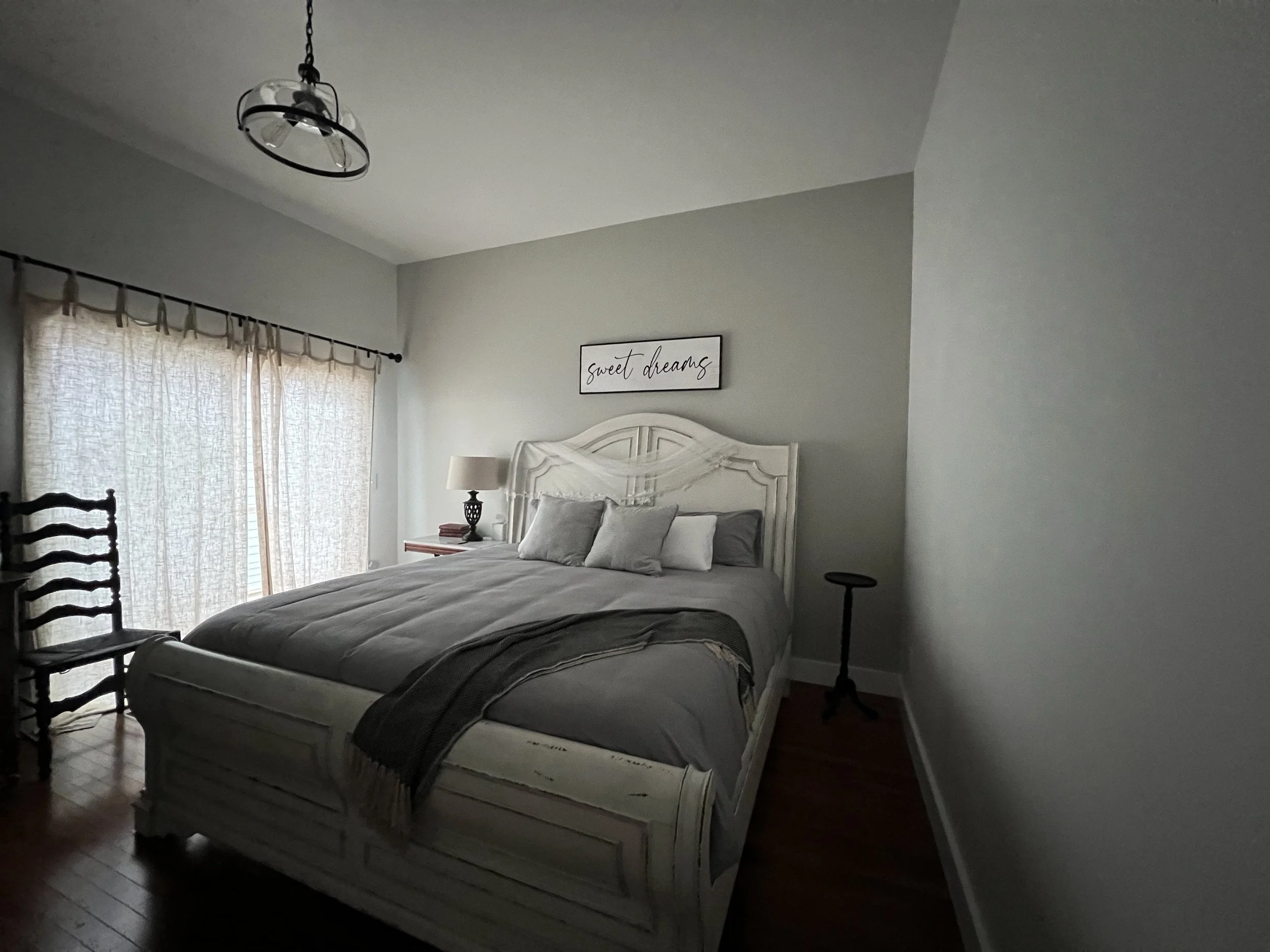A bedroom with a white wooden bed, gray bedding, and multiple gray pillows. A bedside table with a lamp and some books is next to the bed. There is a window with light-colored curtains and a sign on the wall that says "sweet dreams."