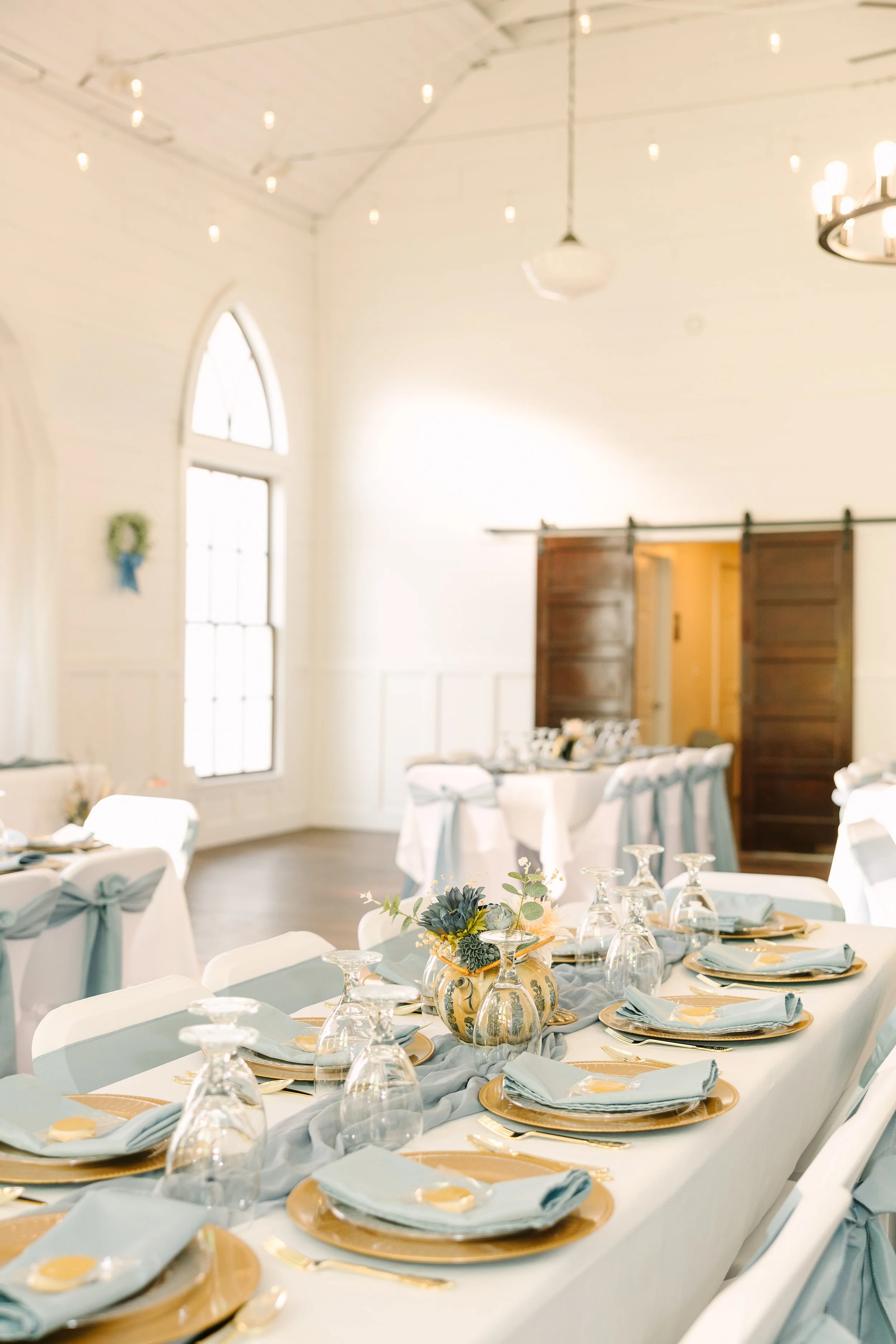 A decorated dining table set for an event in a bright room with white walls and high arched windows, featuring a floral centerpiece, gold-rimmed plates, silverware, and light blue napkins.