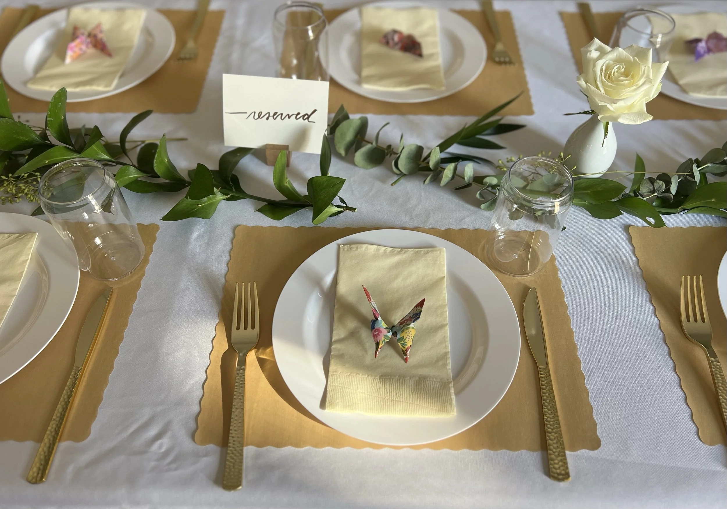 A decorated dining table set for a party, featuring white plates, gold cutlery, glasses, and a floral centerpiece with greenery and a single white rose in a white vase. There are yellow napkins and a small, colorful origami butterfly on one of the pl
