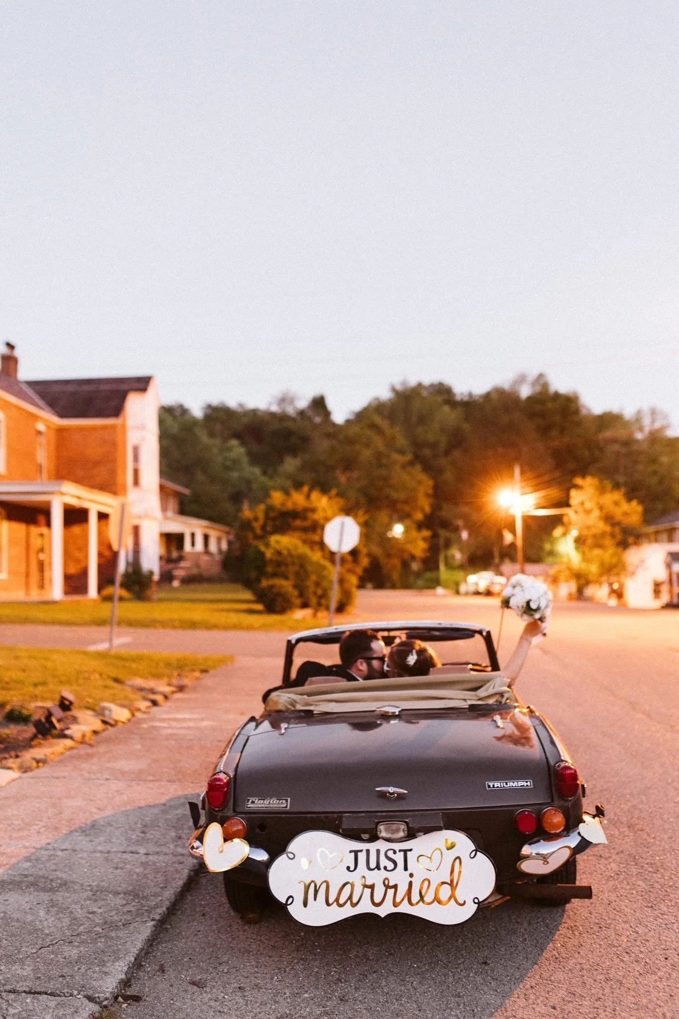 A newlywed couple in a black convertible car decorated for their wedding, with a sign on the back reading 'Just Married.' The bride is holding a bouquet and raising her hand. The scene is at dusk in a residential neighborhood with houses, street ligh