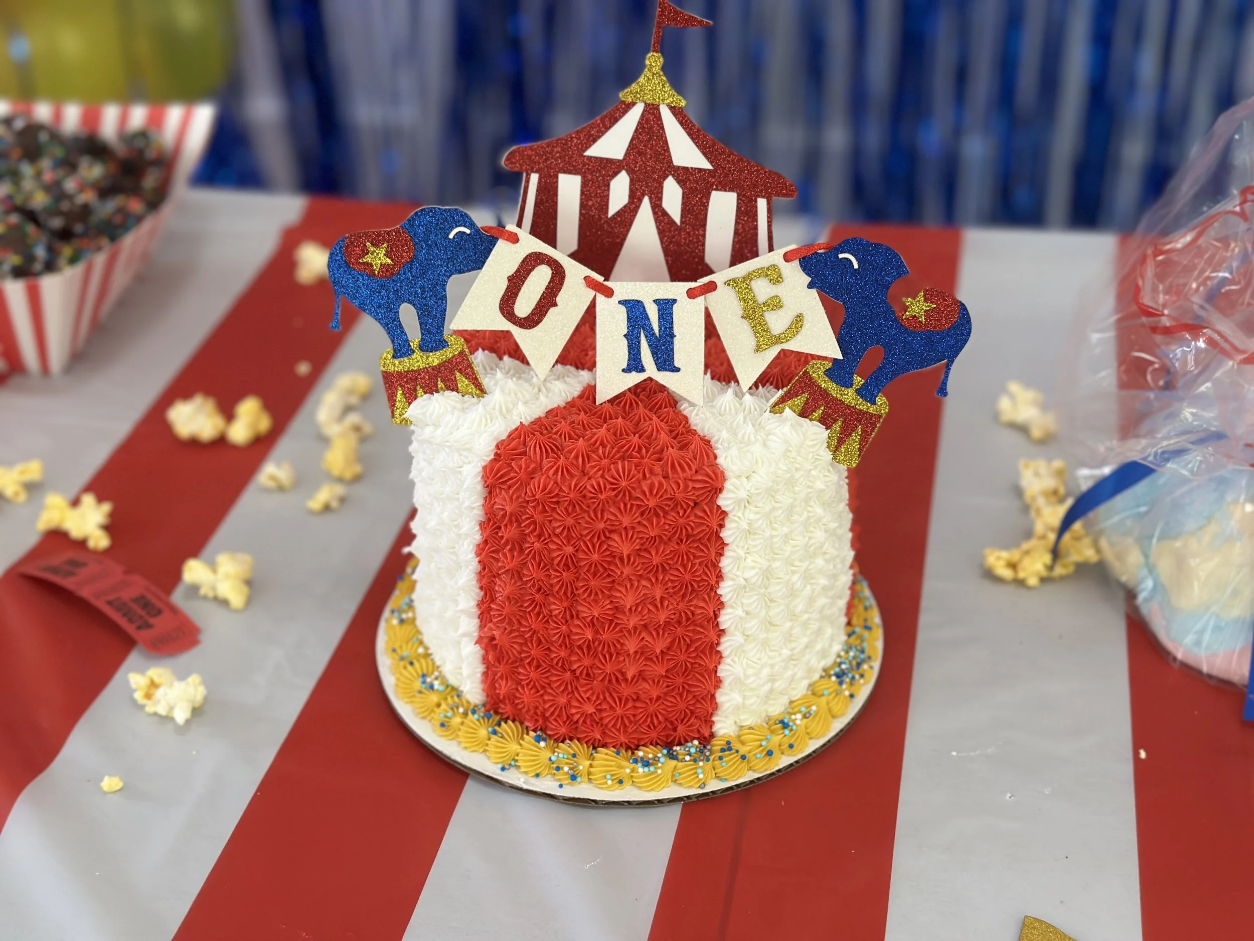 A festive cake decorated with red, white, and blue frosting, topped with a small circus tent, cheerleader figurines, and a banner spelling 'ONE'