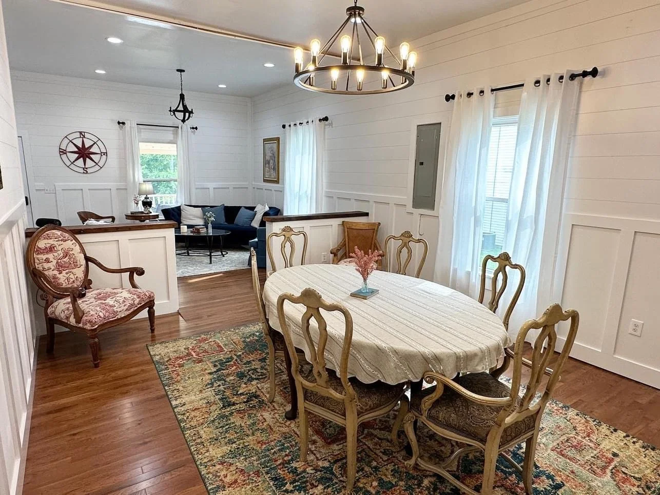Dining room with an oval table, eight vintage-style chairs, and a floral patterned rug. The room has white paneled walls, large windows with white curtains, and a modern chandelier. In the background is a living area with a dark blue sofa, a coffee t