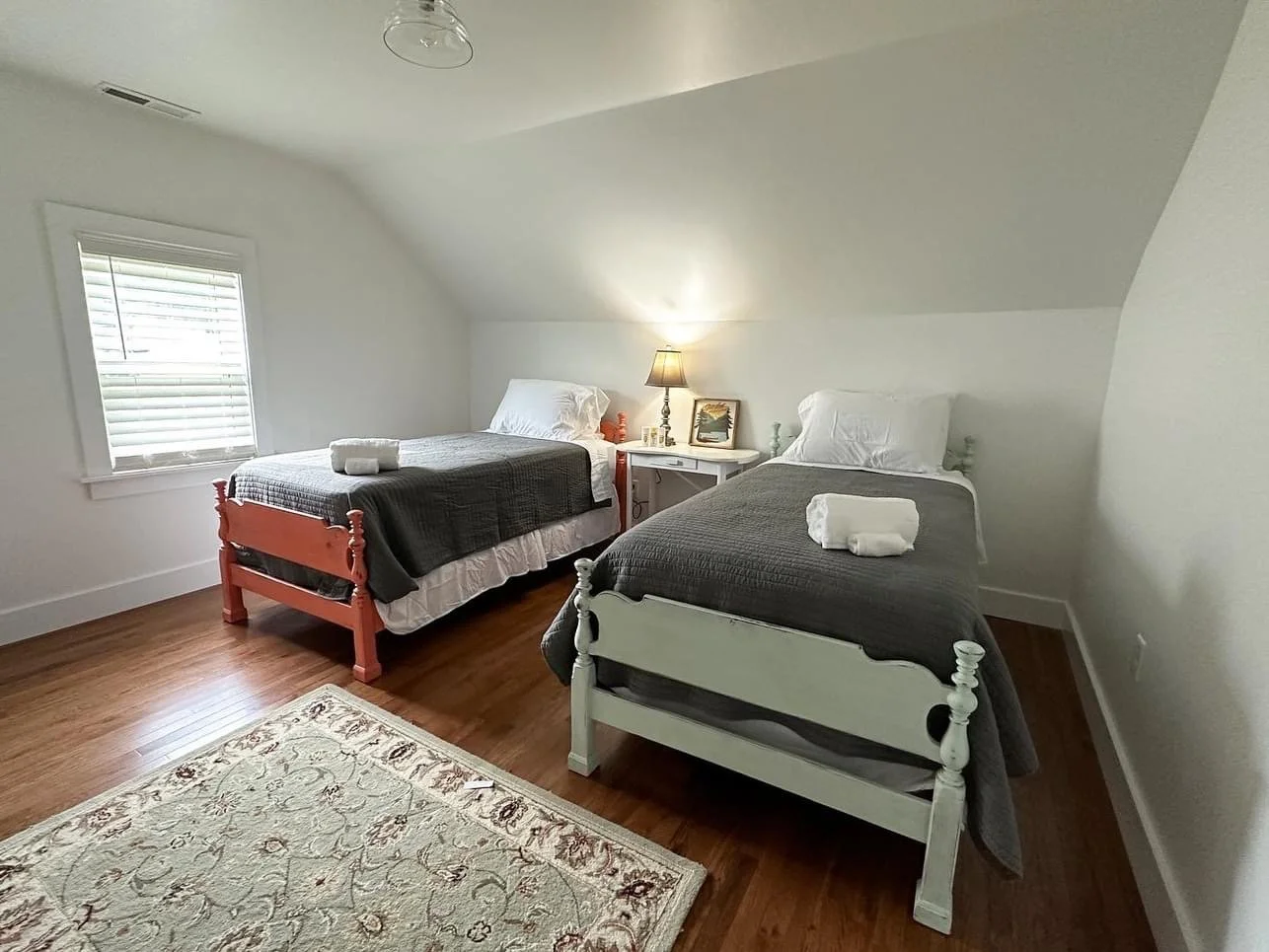 A bedroom with two twin beds, one painted pink and one painted white, with gray bedspreads and towels. There is a window with blinds, a nightstand with a lamp and decor, and a patterned rug on the wooden floor.