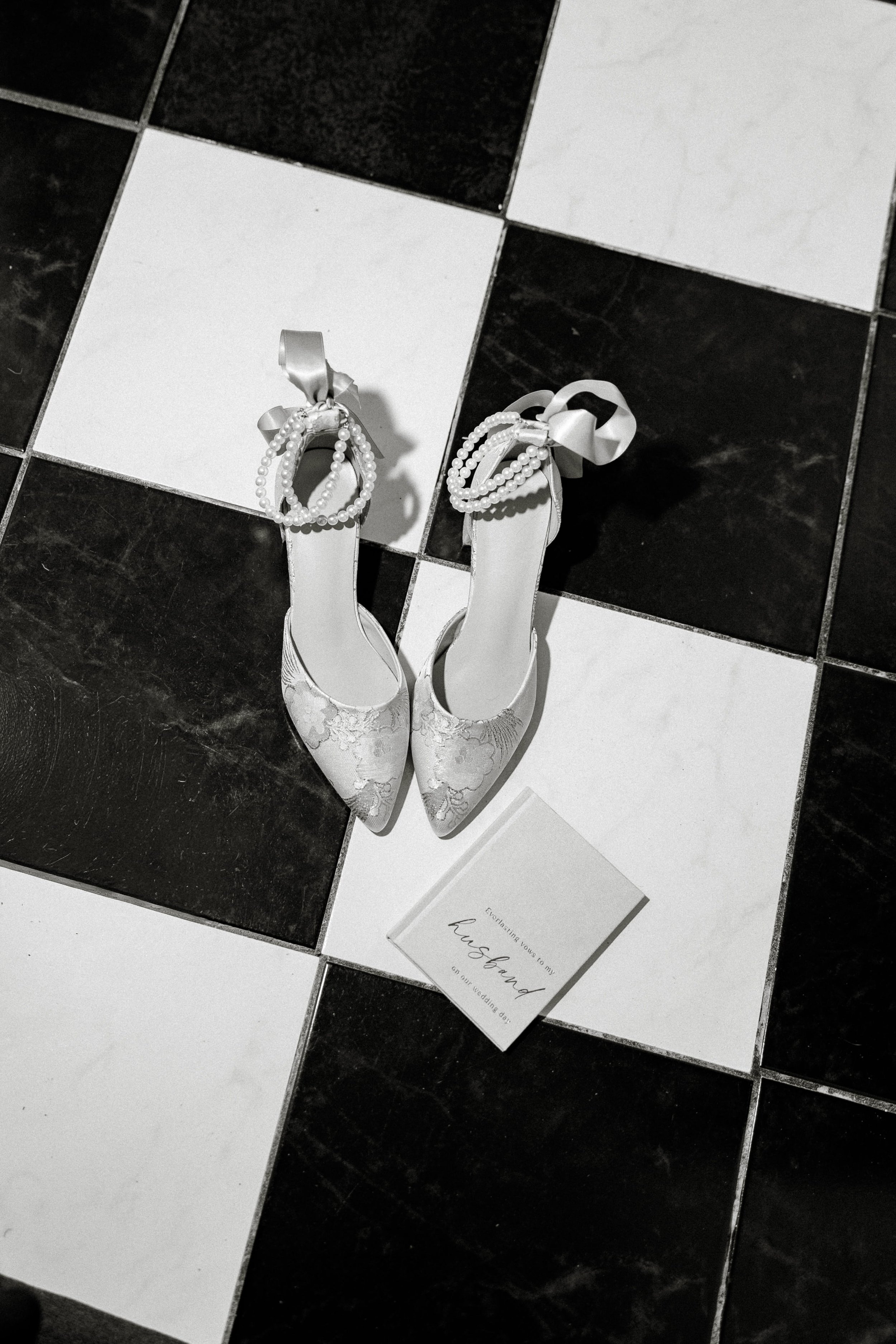 A pair of white lace wedding shoes with decorative bows and pearl embellishments, placed on a black and white tiled floor near a wedding invitation card.