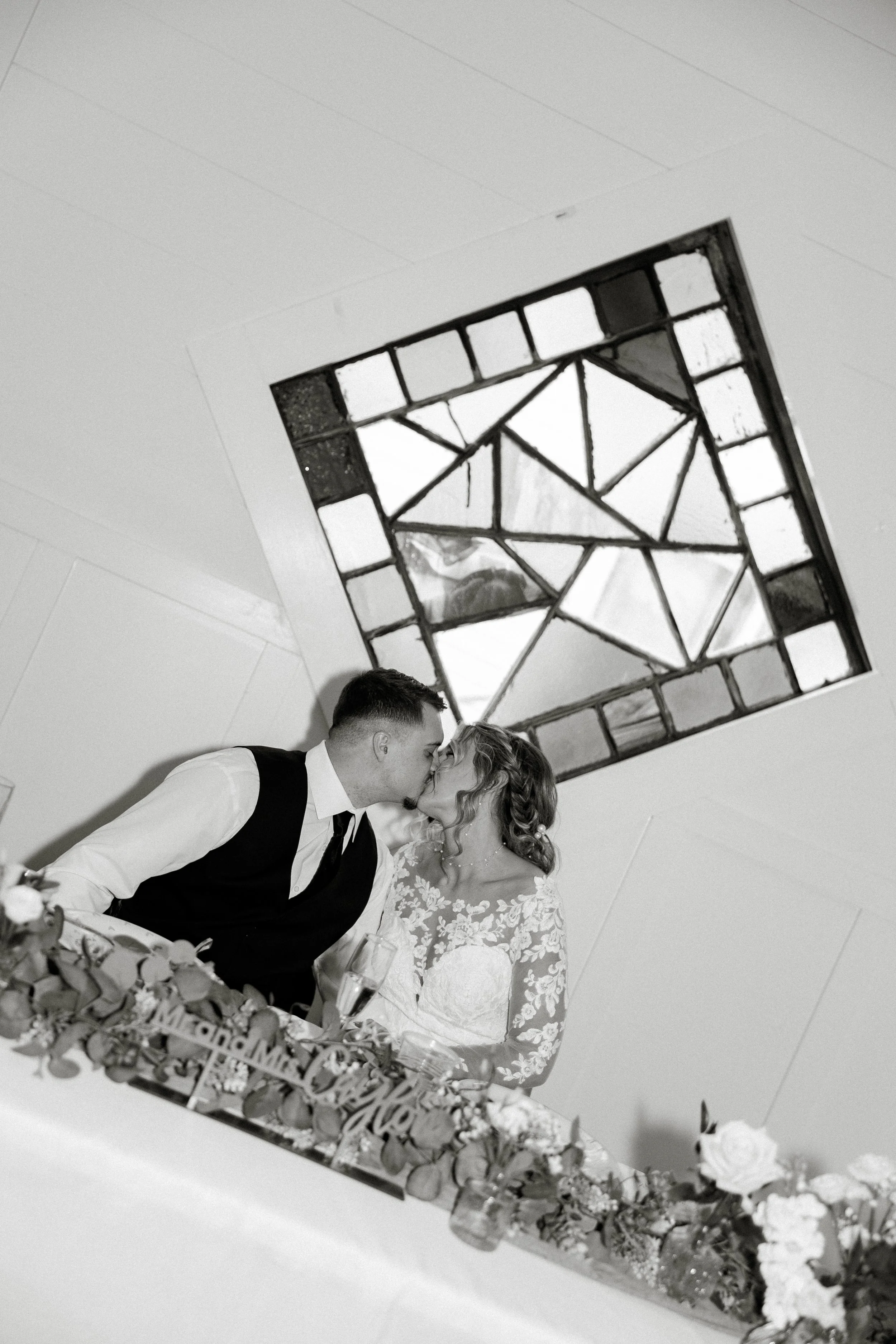 A black and white photo of a wedding scene. The bride and groom are kissing, with the groom dressed in a vest and tie and the bride in a lace wedding dress. They are sitting at a table decorated with flowers and celebrating a wedding.