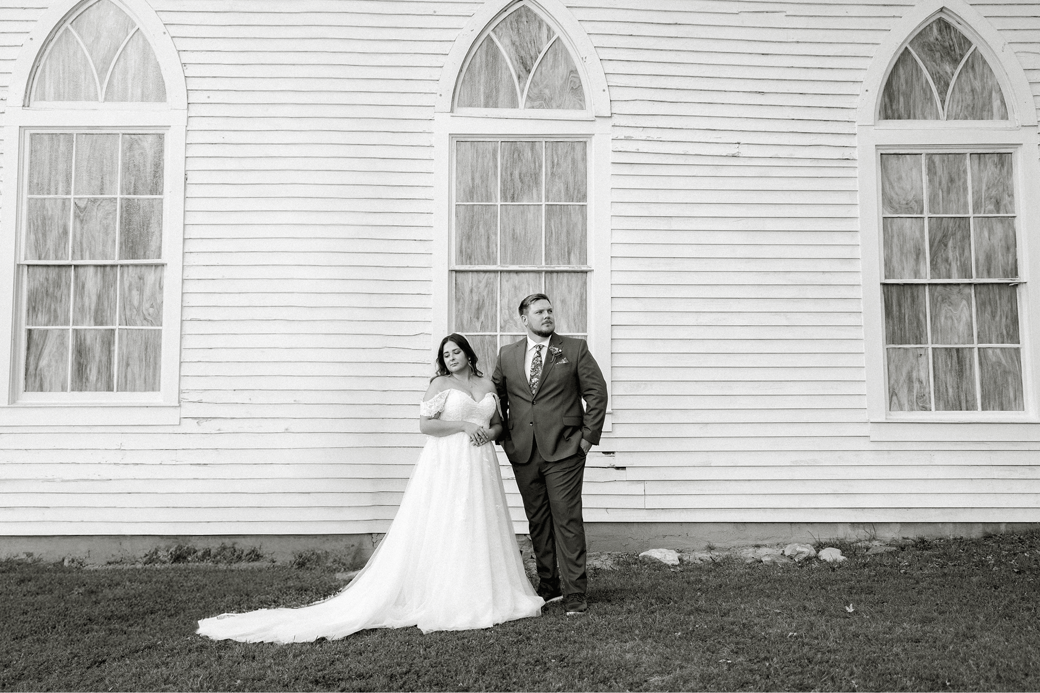 A black and white photo of a bride and groom standing in front of a white wooden building with three large arched windows. The bride is in a white wedding dress and the groom is in a suit with a tie.