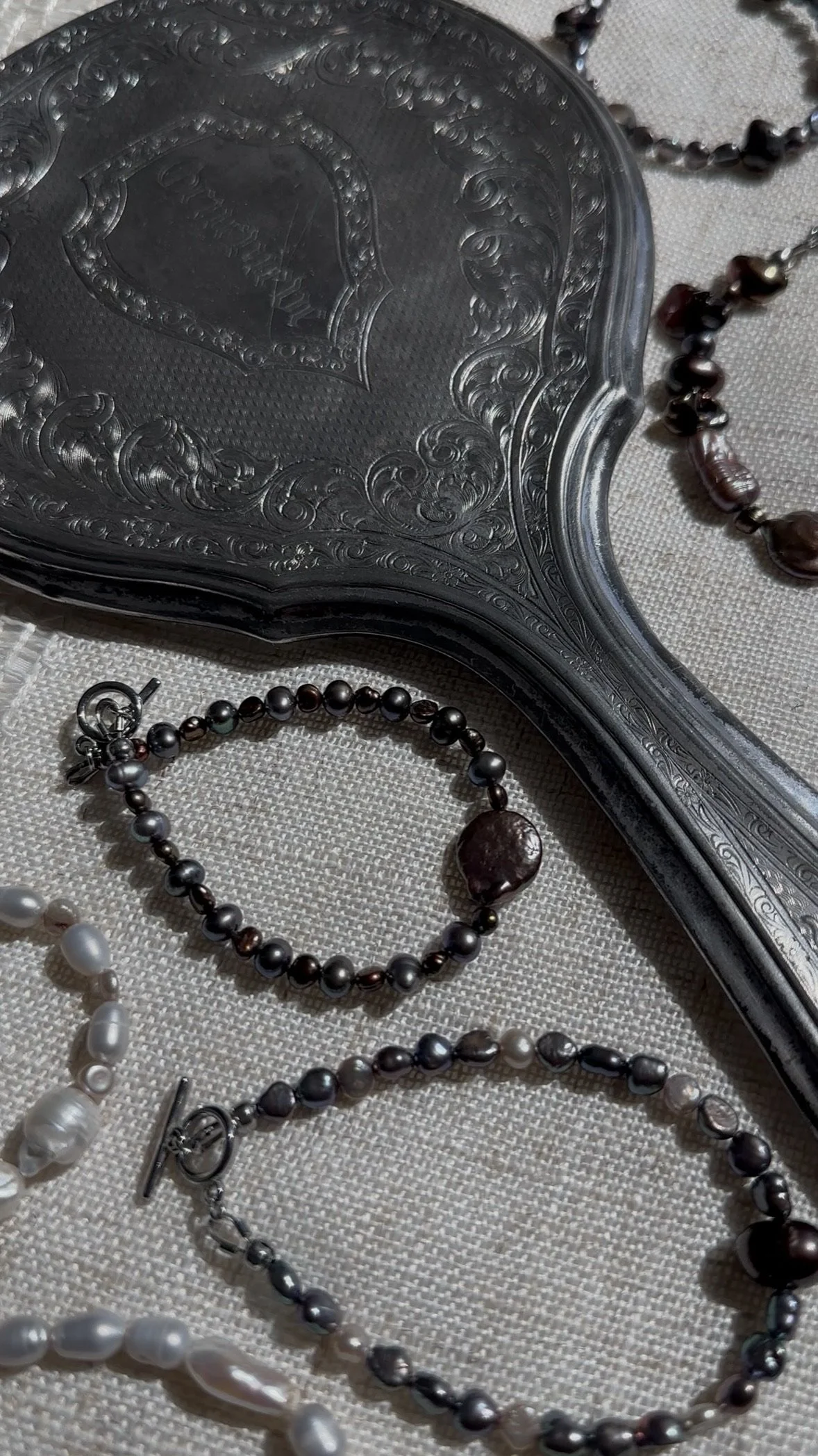 Close-up of a vintage silver hand mirror and several pearl necklaces placed on a textured fabric surface.