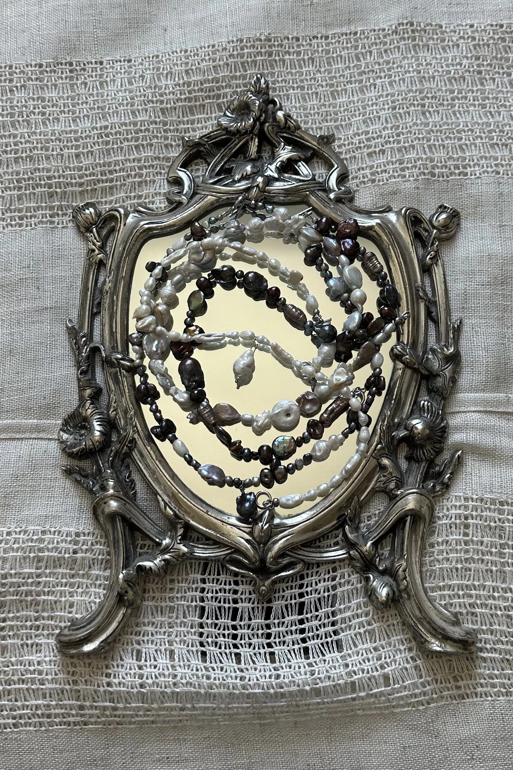 An ornate vintage silver mirror with a decorative frame, lying on a textured fabric surface. Several pearl necklaces, along with other beaded jewelry, are draped over the mirror, partially reflecting in the glass.