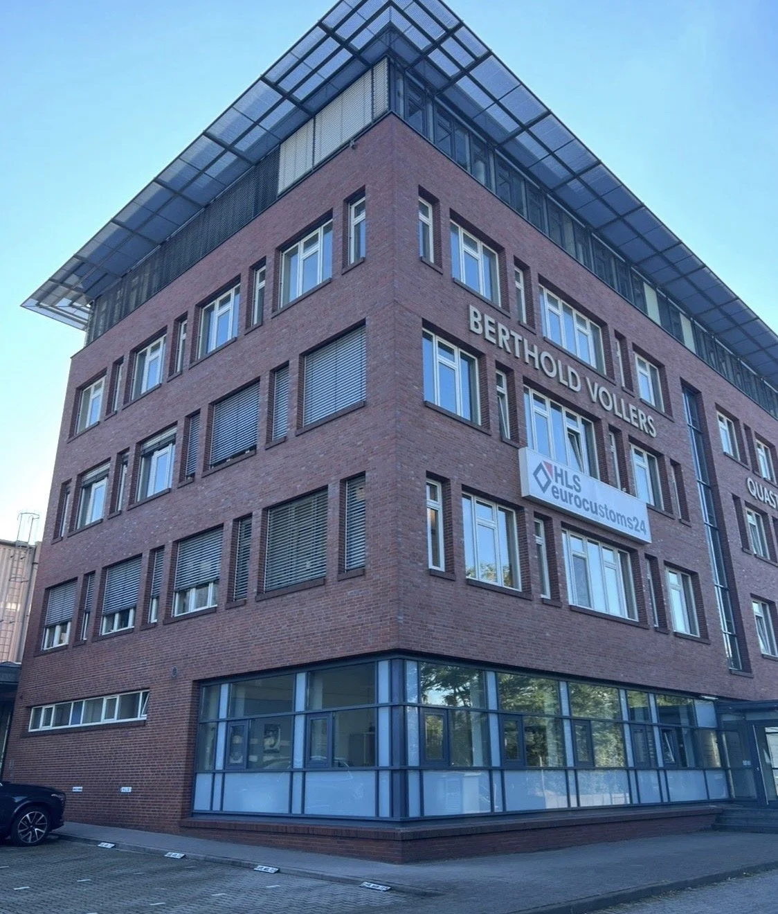 Mehrstöckiges Bürogebäude mit roten Ziegelwänden und großen Fenstern, Beschriftung "BERTHOLD VOLBERS" und einem Schild "HLS Euro Customs 24" auf der Fassade, bei Sonnenschein, Blick nach oben