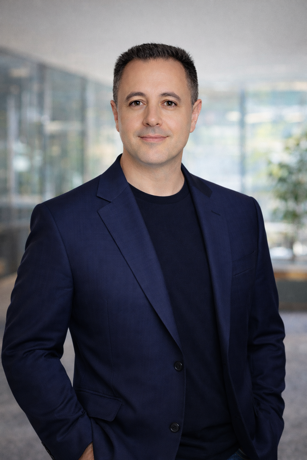 Professional portrait of a man in a navy blazer, standing in a modern office building with glass walls and blurred background.