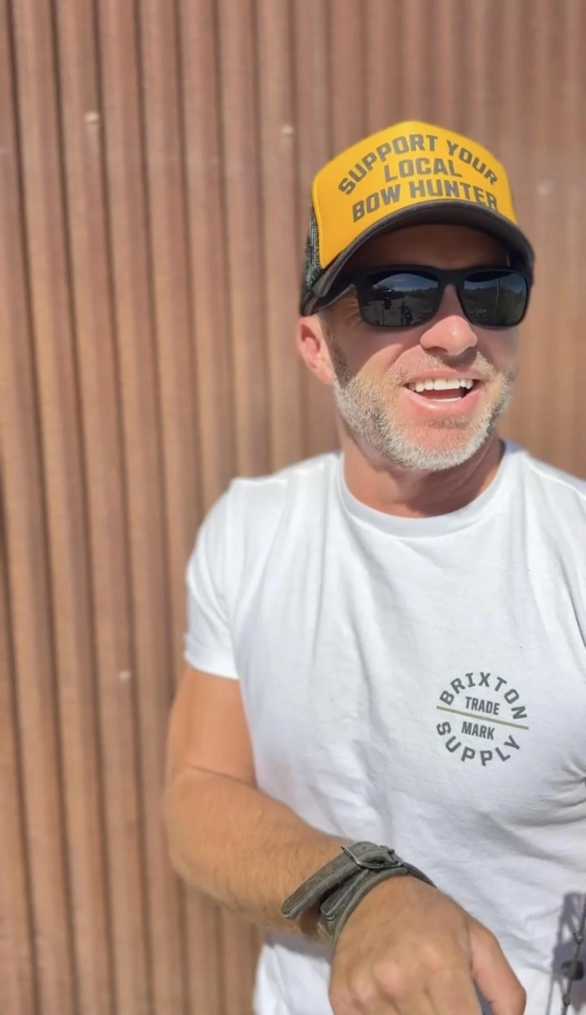 Adam Greentree wearing a white t-shirt, black sunglasses, and a hat  with a yellow front panel that says "Support Your Local Bow Hunter," is smiling and standing in front of a wooden wall.