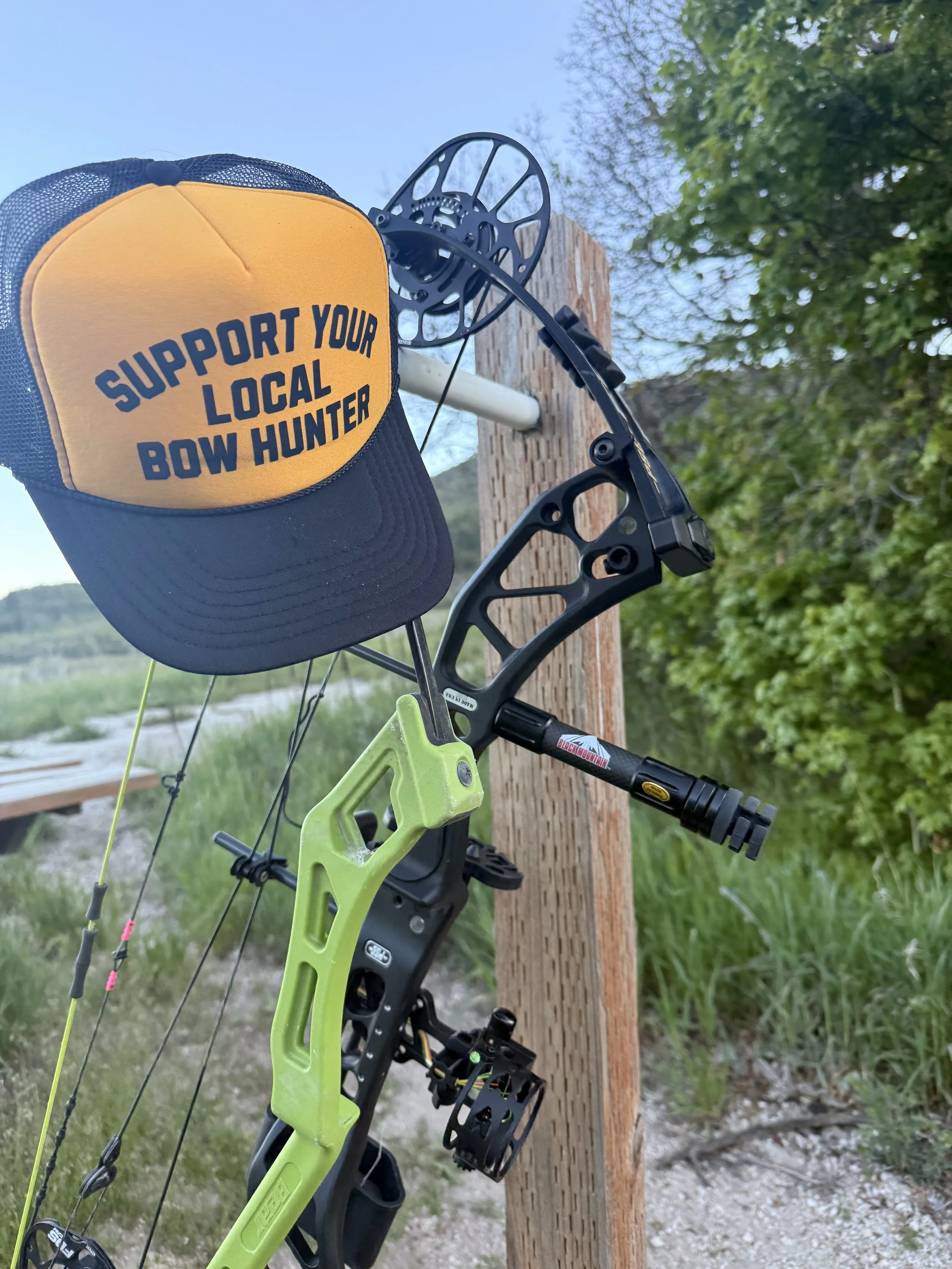 Close-up of a yellow and black cap with the text "Support your local bow hunter" hung on the string of a green compound bow. The bow is mounted on a wooden post outdoors with green trees and sky in the background.