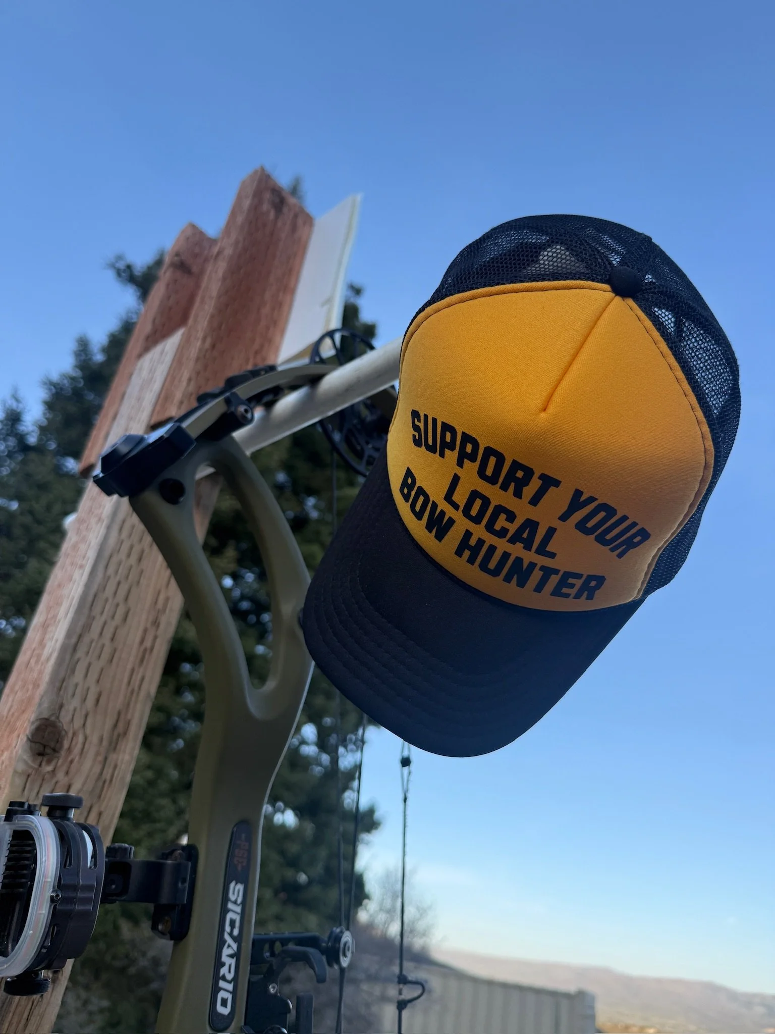 Close-up of a yellow and black support gear labeled 'Support Your Local Bow Hunter' hanging on a bicycle handlebar next to a wooden post, with a blurred outdoor landscape and clear blue sky in the background.