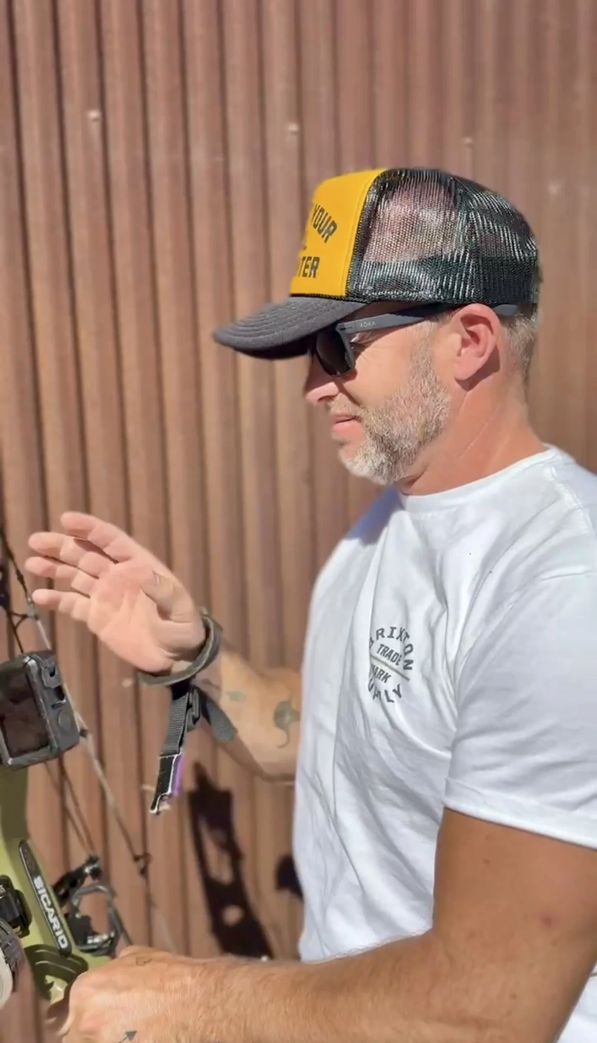 Adam Greentree wearing a Jacey Loren trucker hat, sunglasses, and a white t-shirt looks at a bow while standing in front of a wooden wall.