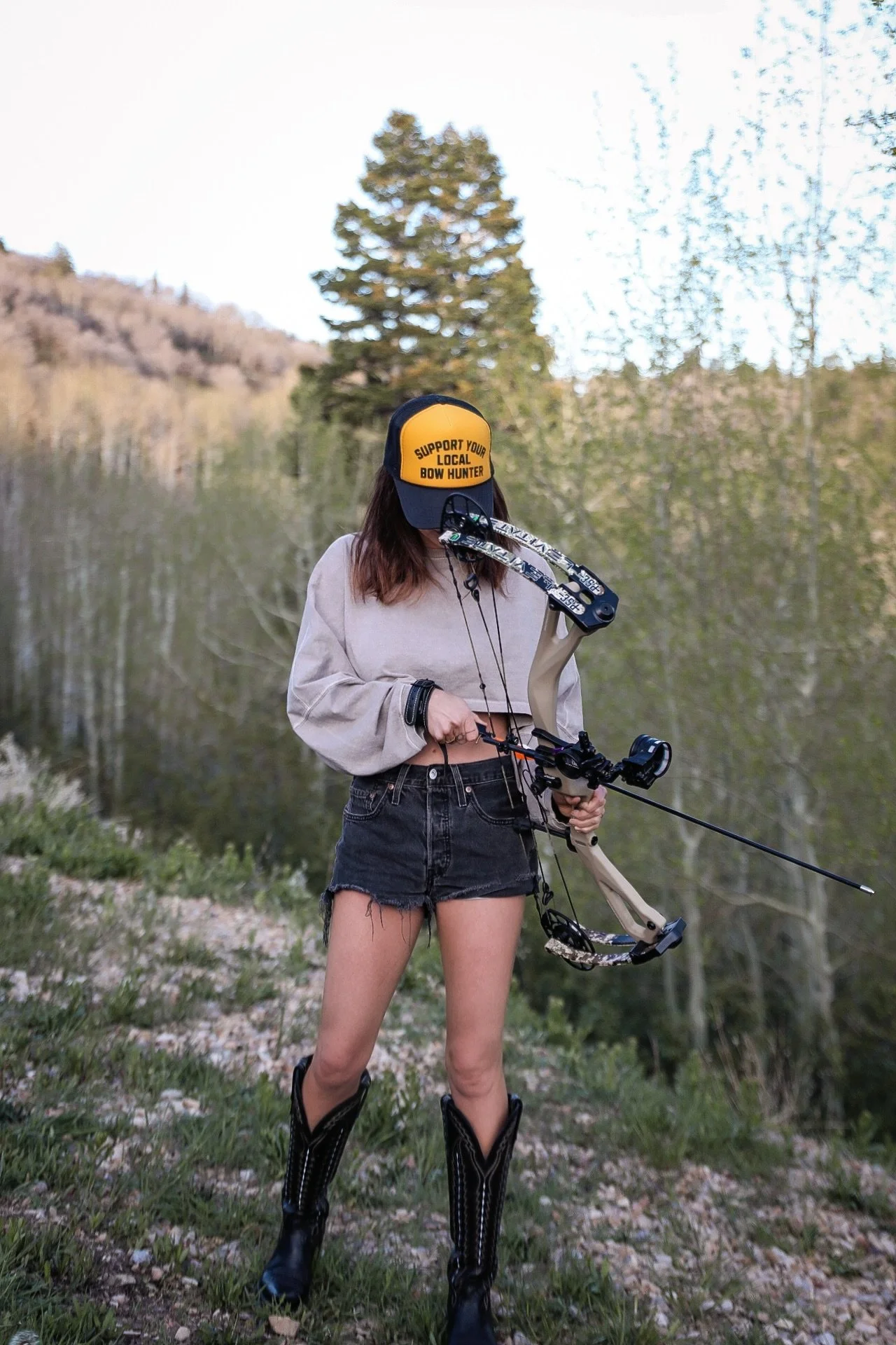 A person with long brown hair wearing a yellow and black hat with words supporting local bow hunters, a beige cropped sweatshirt, black shorts, and black boots holding a compound bow near a body of water, with trees and hills in the background.
