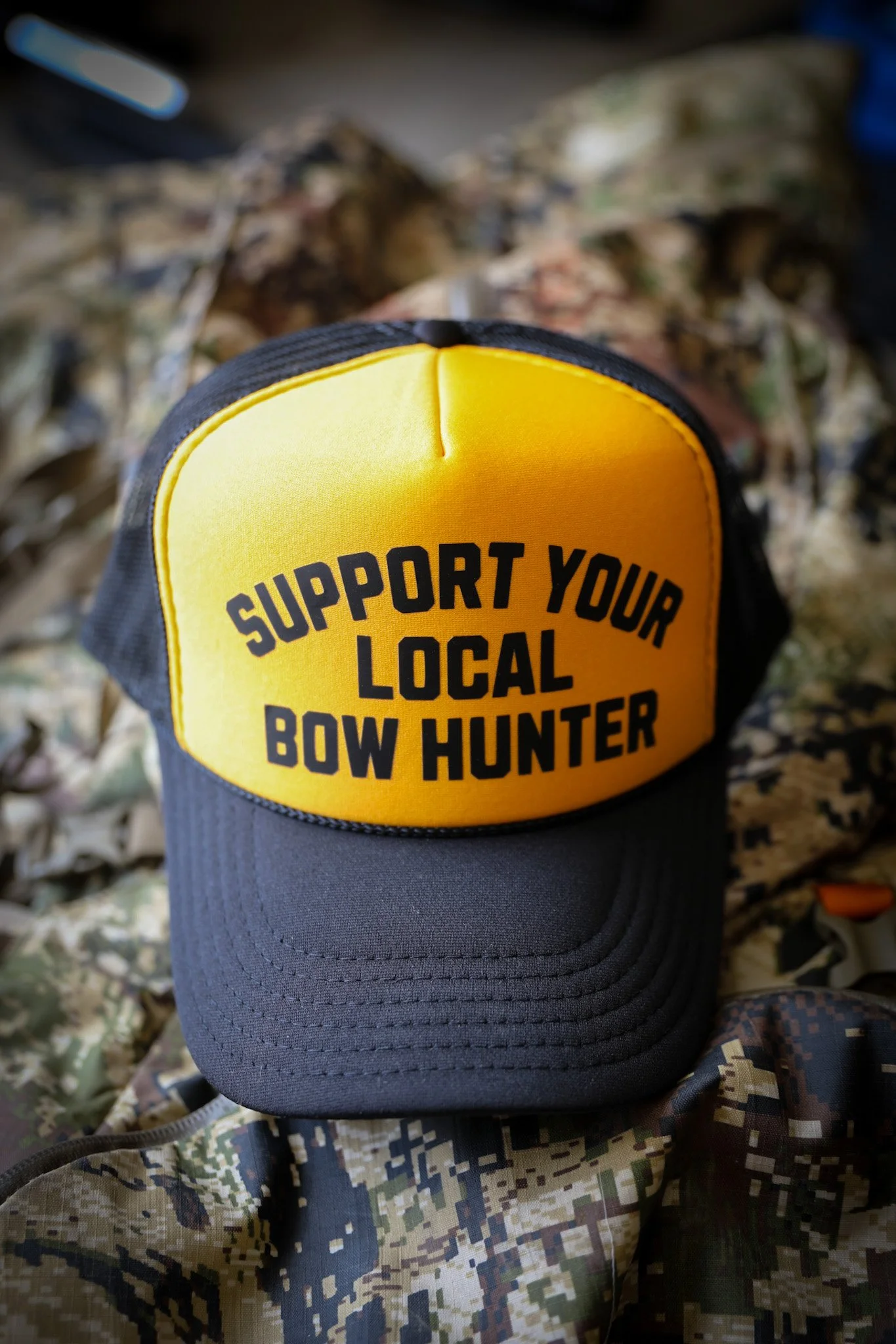 A close-up of a black and yellow hat with the words "Support Your Local Bow Hunter" embroidered on the front, resting on camouflage-patterned fabric.