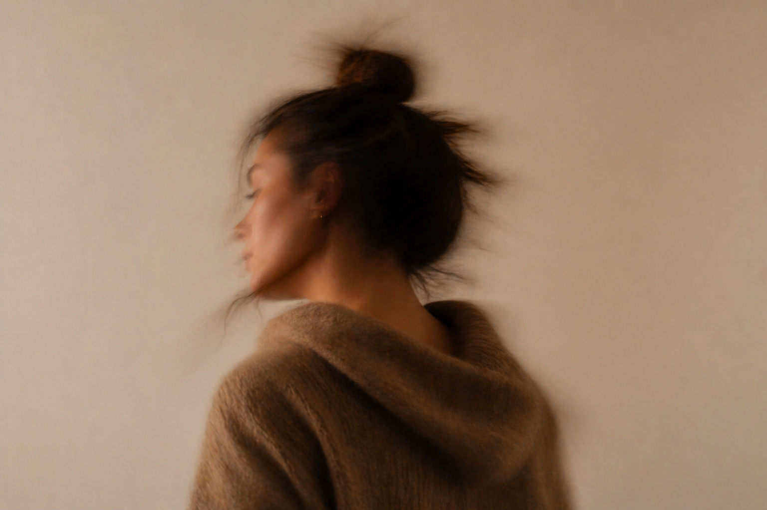 Side profile of a woman with hair tied in a bun wearing a brown sweater, blurred background.
