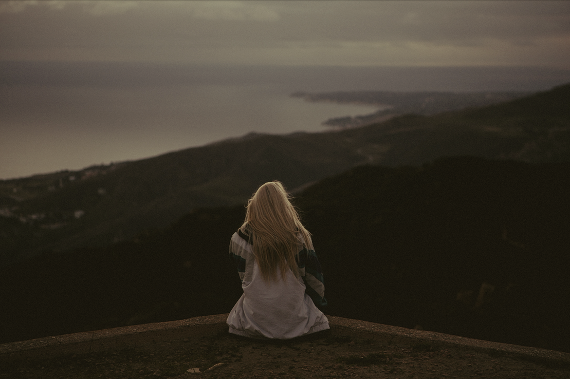 A woman with long blonde hair sits on a ledge overlooking a hilly landscape and water at dusk, with dark clouds overhead.