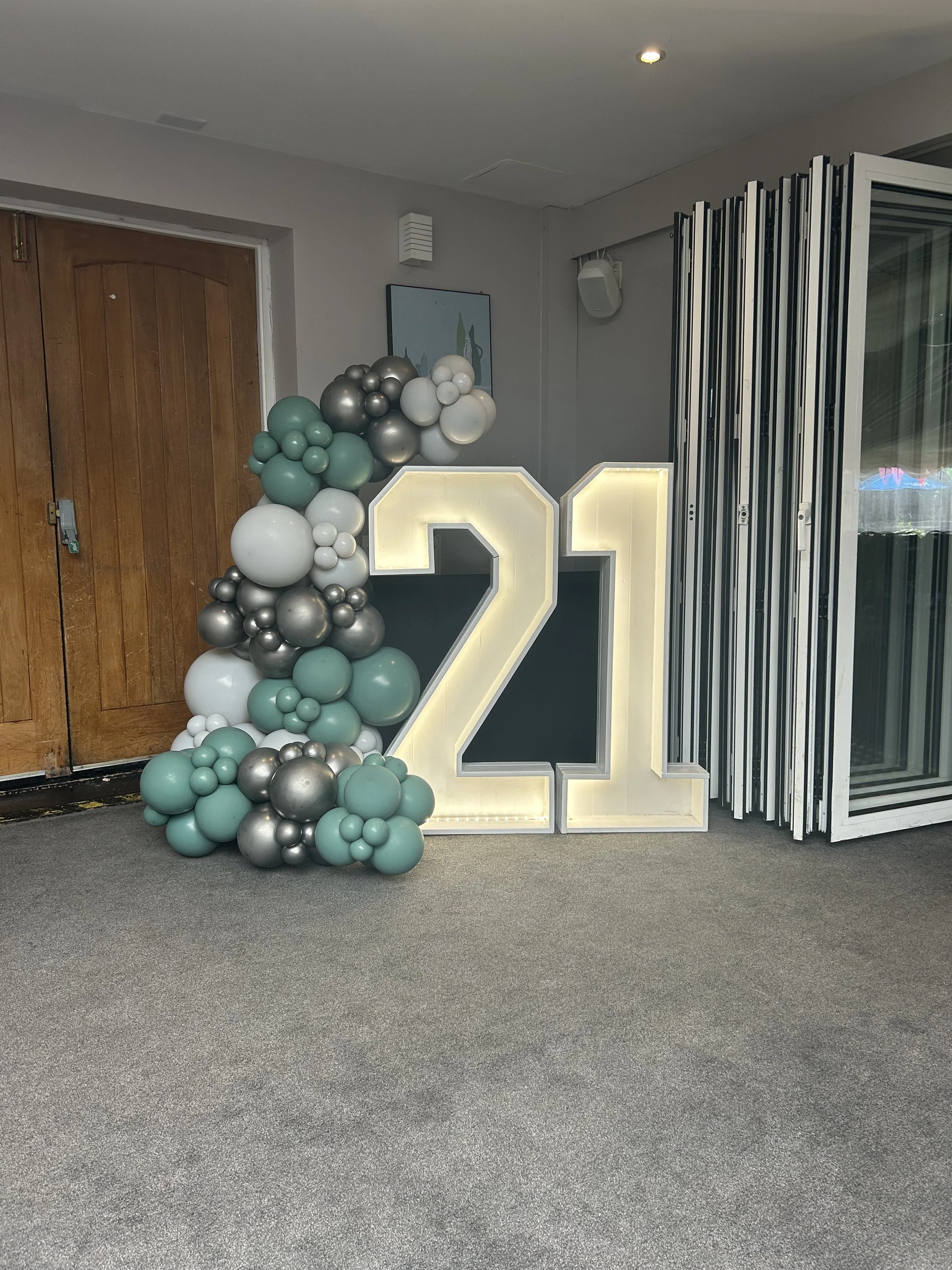 Decorative display with a large illuminated number two and four, a cluster of balloons in shades of teal, gray, black, and white, set against a gray wall with a wooden door and modern decor.