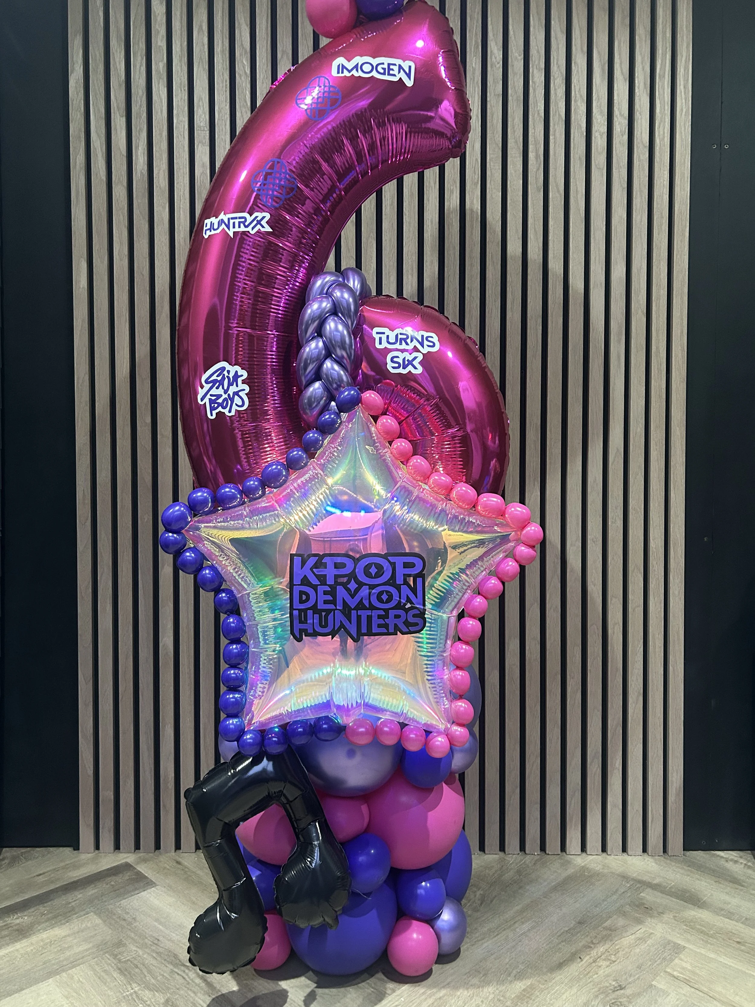 A colorful balloon display with pink, purple, and black balloons, including a large pink number 6 balloon and a star-shaped iridescent balloon with the words 'KPOP DEMON HUNTERS'.