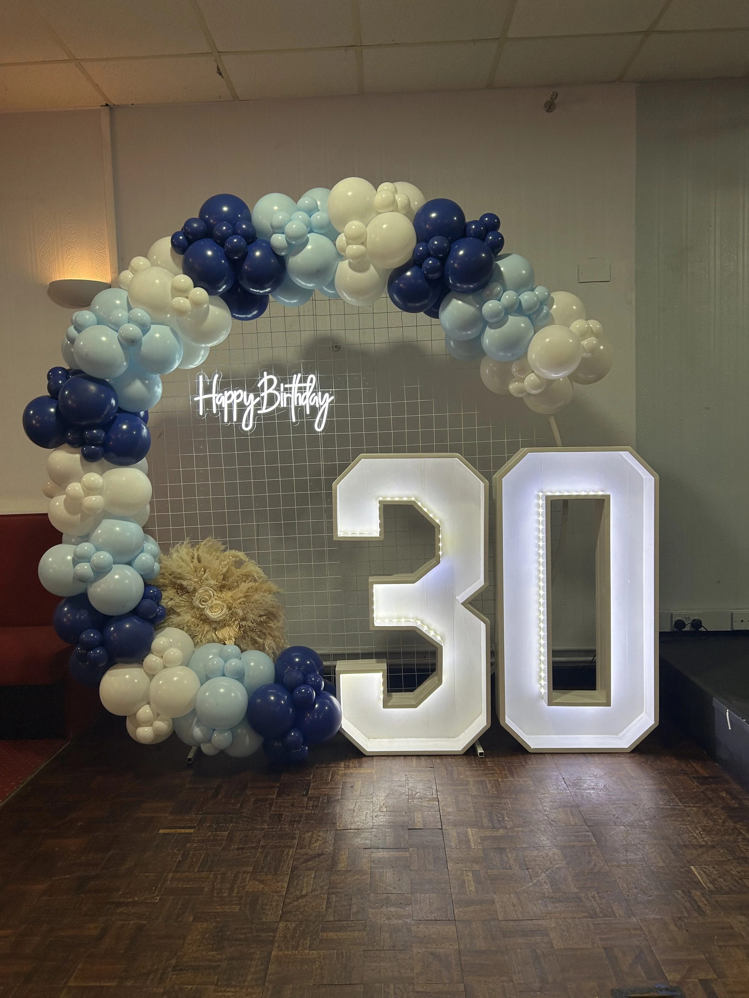 Illuminated large white number '30' with balloon arch in shades of blue, white, and beige, and a neon sign that says 'Happy Birthday' against a gray tiled wall.