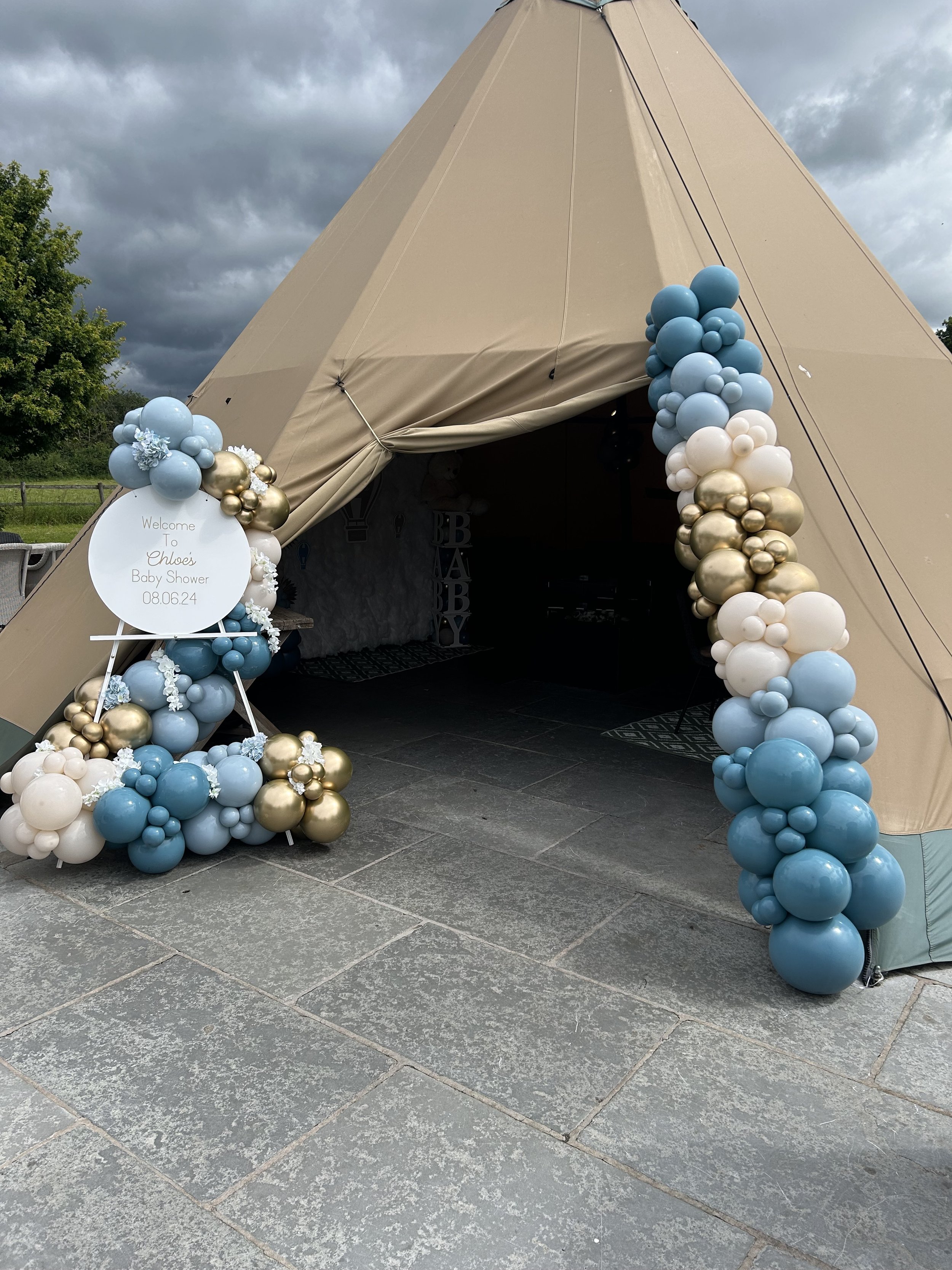 A beige teepee decorated with blue, white, and gold balloons and floral accents at the entrance, with a sign indicating a baby shower for Chloe on August 6, 2024.